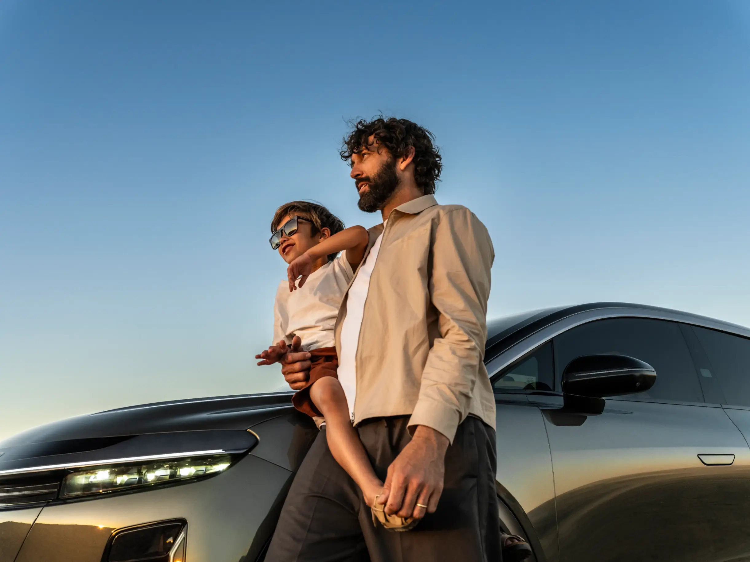Die Aufnahme zeigt einen Mann, der vor einem Lucid Air Pure in Infinite Black Metallic auf die Kamera zugeht. Der Lucid Air ist mit Dachgepäckträger ausgestattet, der mit einem Kanu beladen ist. Im Hintergrund sind ein See, Bäume und hohe, felsige Berge zu sehen.