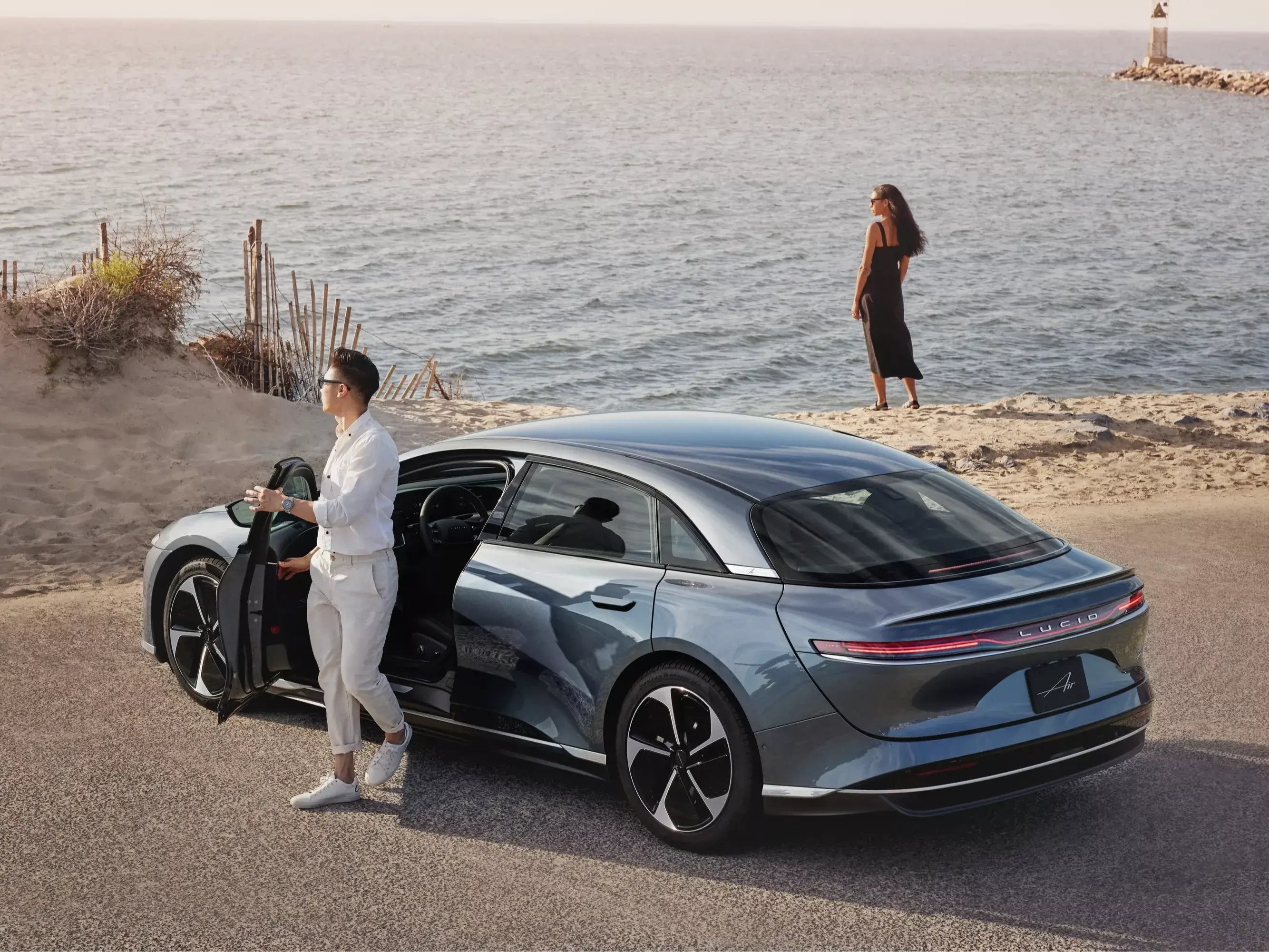 Die Aufnahme zeigt einen Mann, der vor einem Lucid Air Pure in Infinite Black Metallic auf die Kamera zugeht. Der Lucid Air ist mit Dachgepäckträger ausgestattet, der mit einem Kanu beladen ist. Im Hintergrund sind ein See, Bäume und hohe, felsige Berge zu sehen.