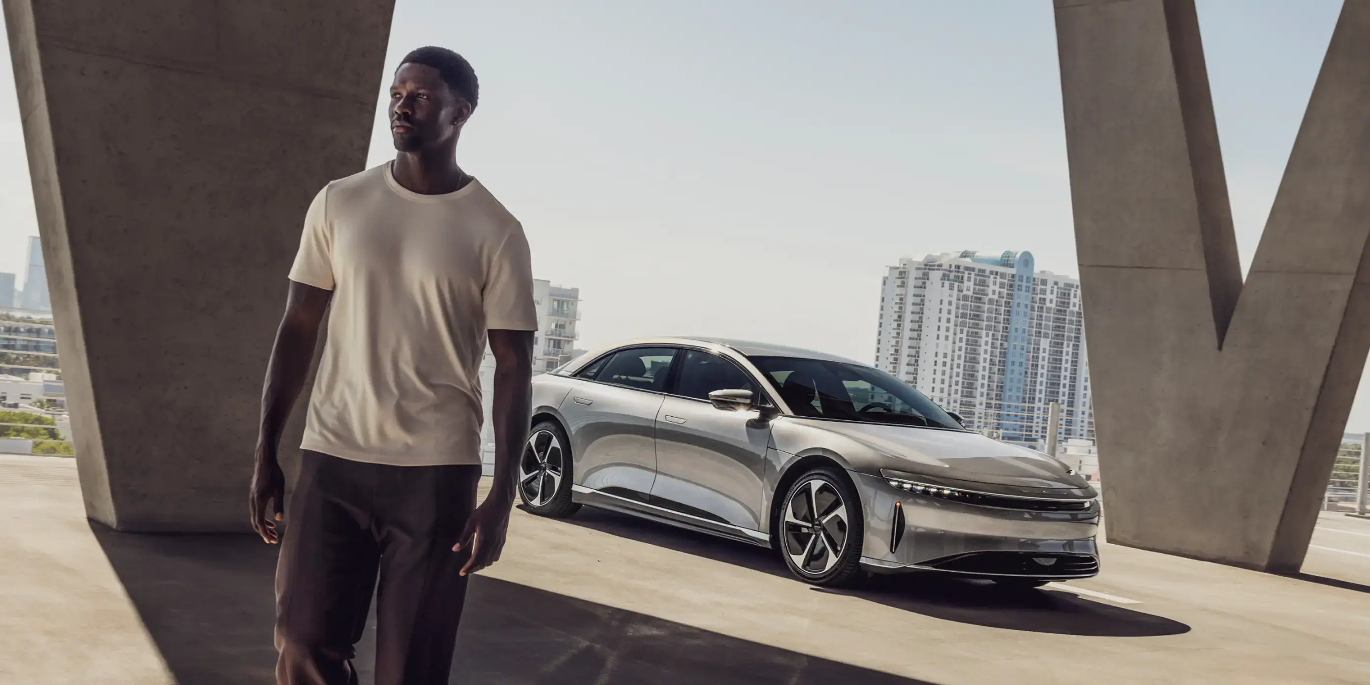 رجل يسير نحو الكاميرا أمام سيارة Lucid Air Pure باللون الأسود إنفينيت المعدني. سيارة Lucid Air مجهزة بحامل فوق السقف يحمل زروق كنو. وفي الخلفية تظهر بحيرة وأشجار وسلسلة طويلة من الجبال.