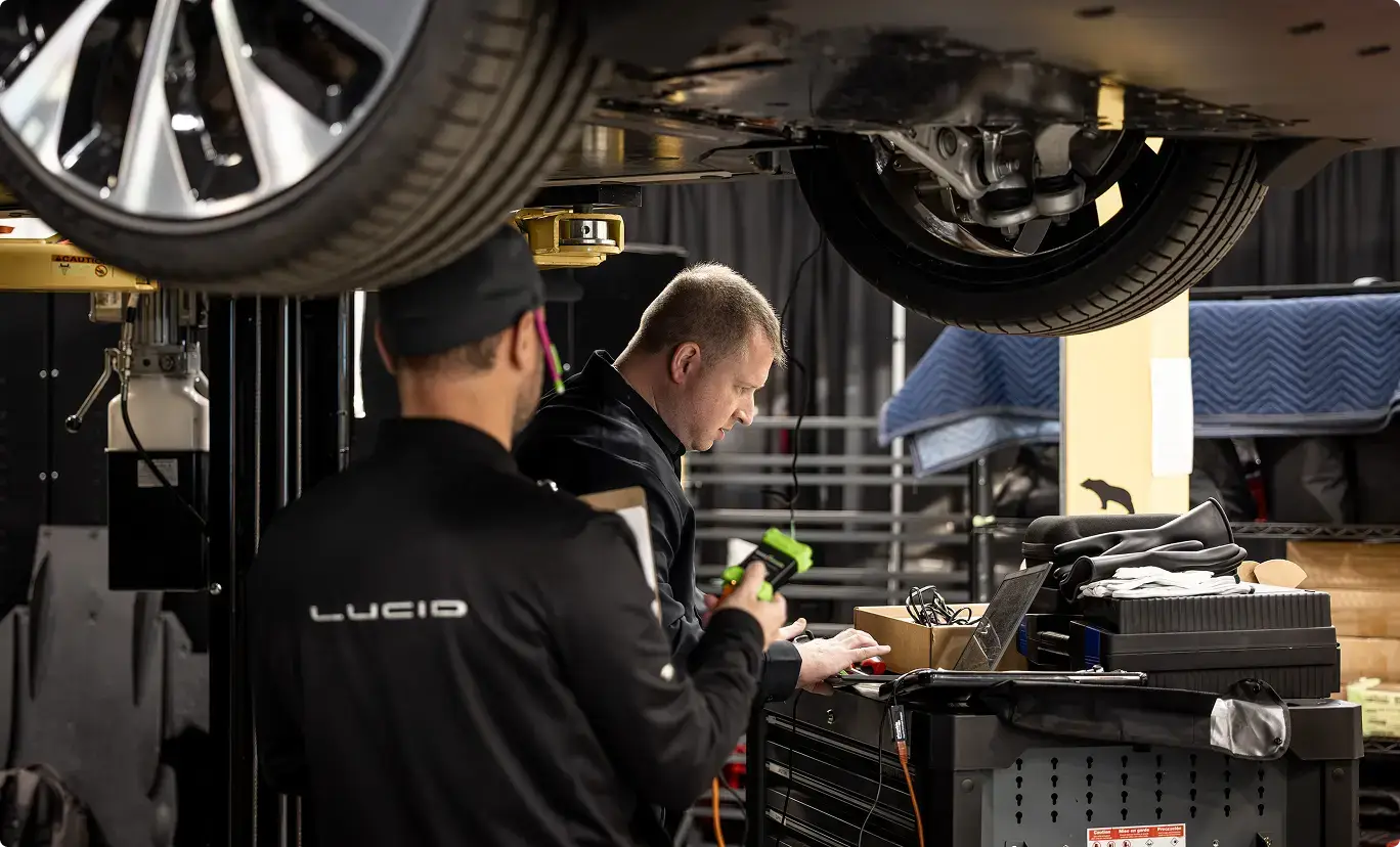 Mechanics working beneath a vehicle on a lift in an auto shop, with tools and equipment visible on a workbench.