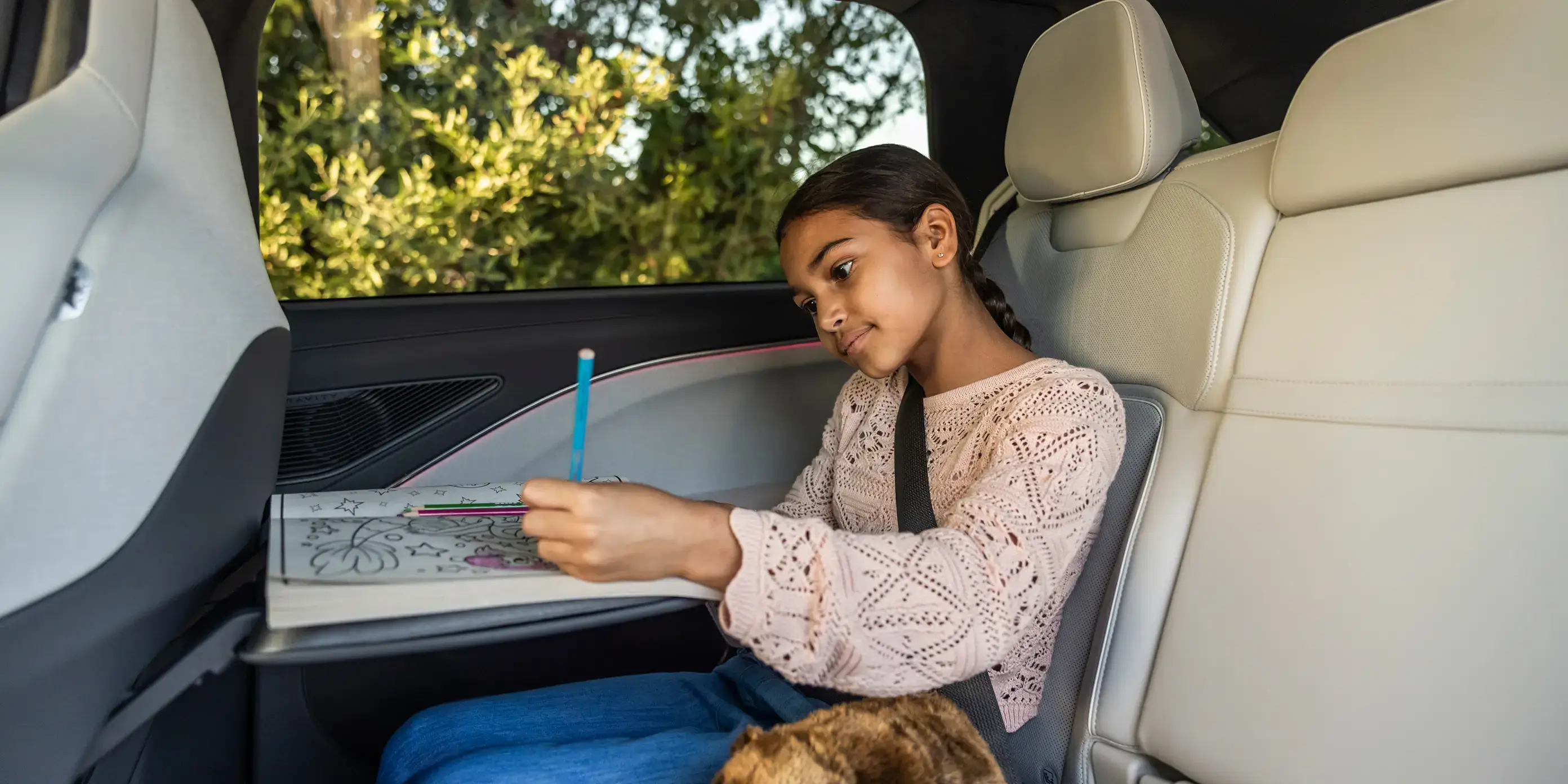 Child in Lucid Gravity backseat colouring in a book while wearing a seatbelt, with cream interior and greenery visible outside.