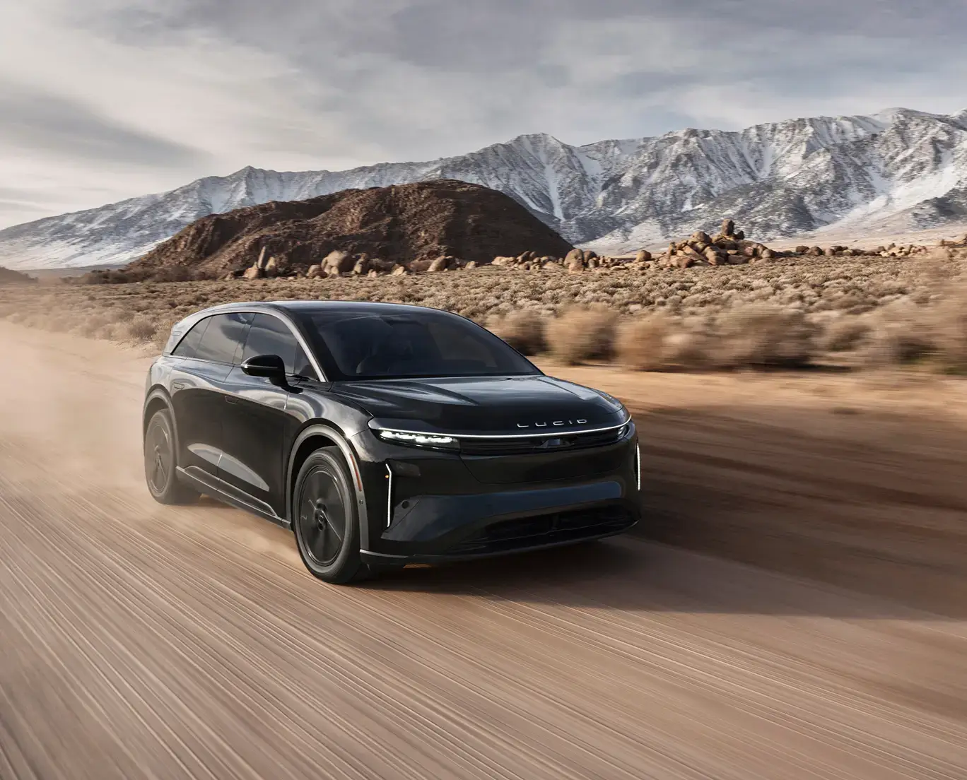 Black Lucid electric SUV driving on desert road with snow-capped mountains in background under cloudy sky.
