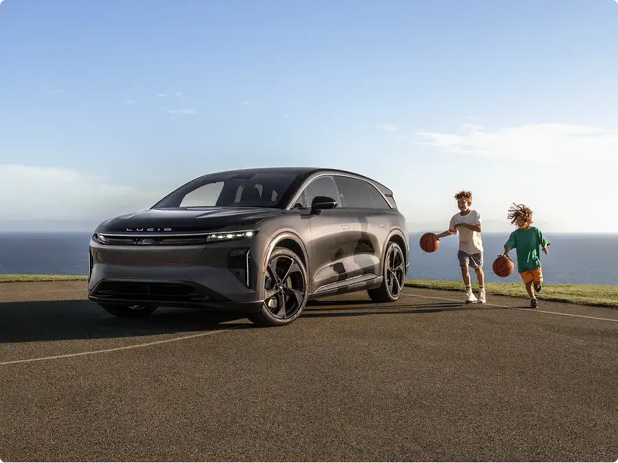 A Lucid SUV parked outdoors near the ocean, with two children dribbling basketballs beside it under a clear sky.