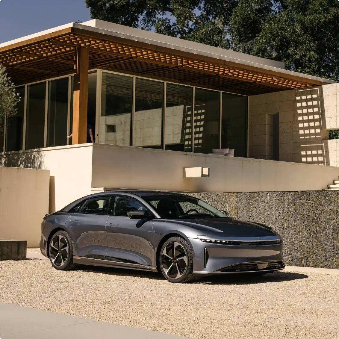 A sleek gray luxury electric sedan parked on a gravel driveway in front of a modern architectural home.