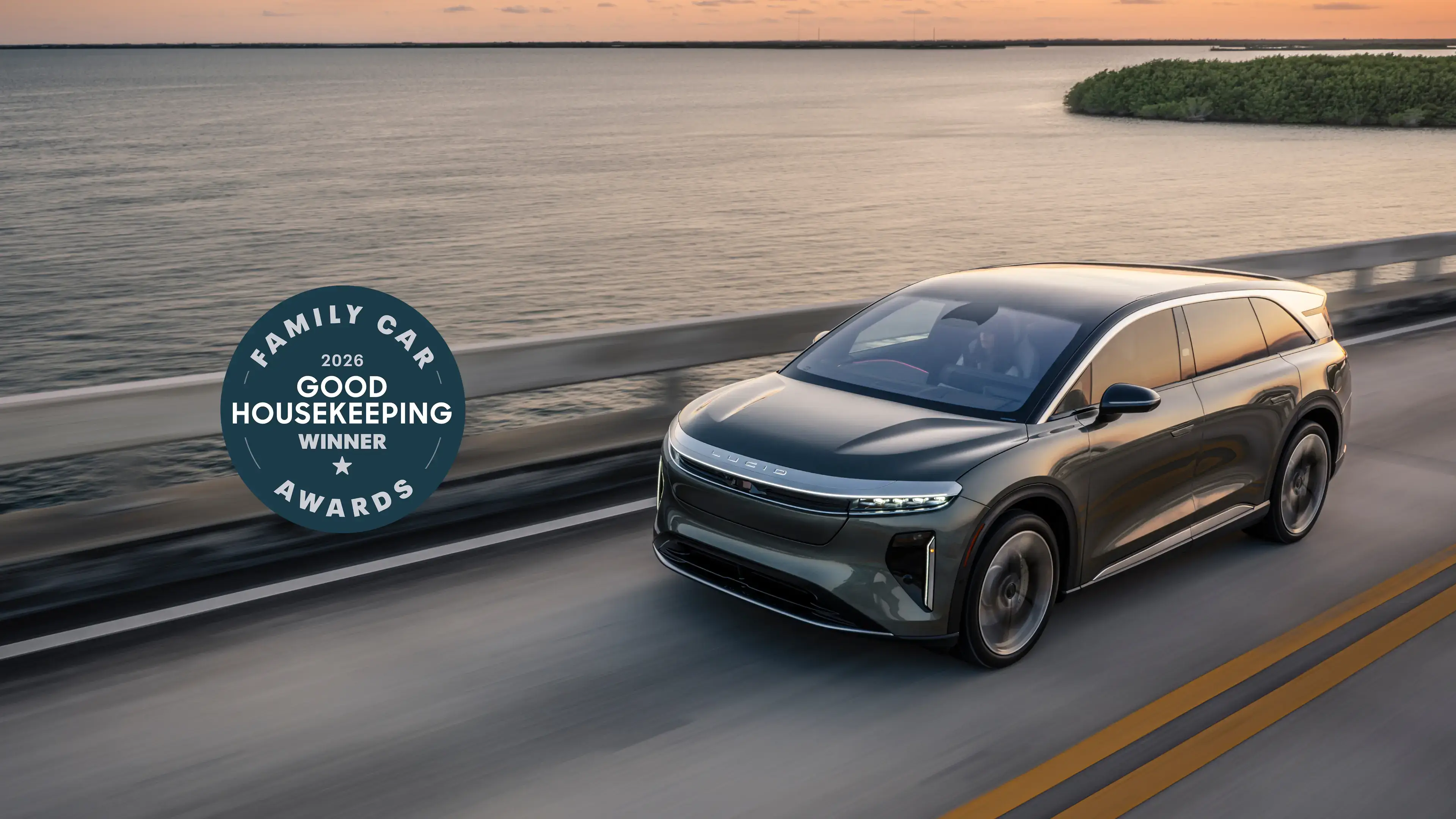 Lucid Gravity driving along coastal highway at sunset, featuring Good Housekeeping 2026 Family Car Award winner badge.
