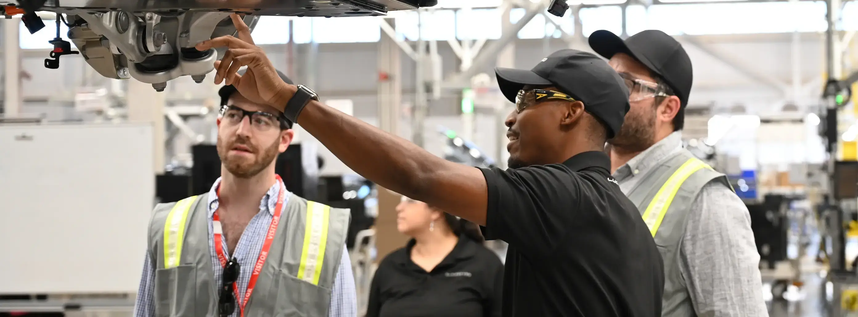 Automotive technicians in safety gear examining the underside of a vehicle on an assembly line in a factory setting.