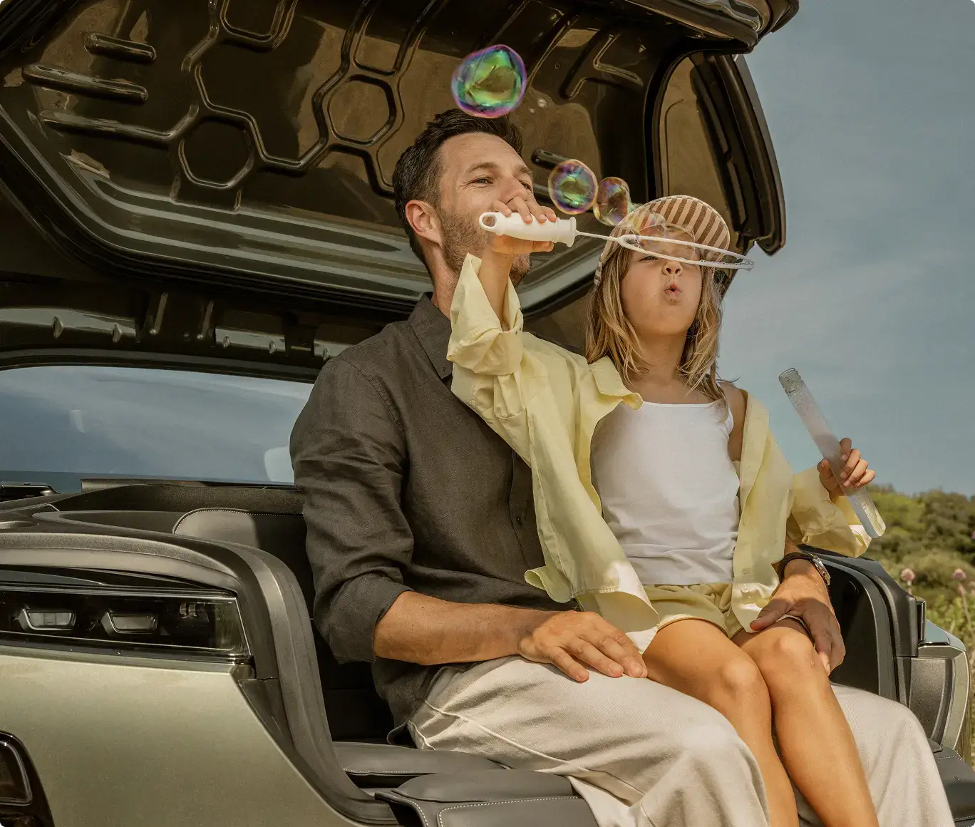 A man and young child sit in an open car trunk blowing colorful soap bubbles together on a sunny day.