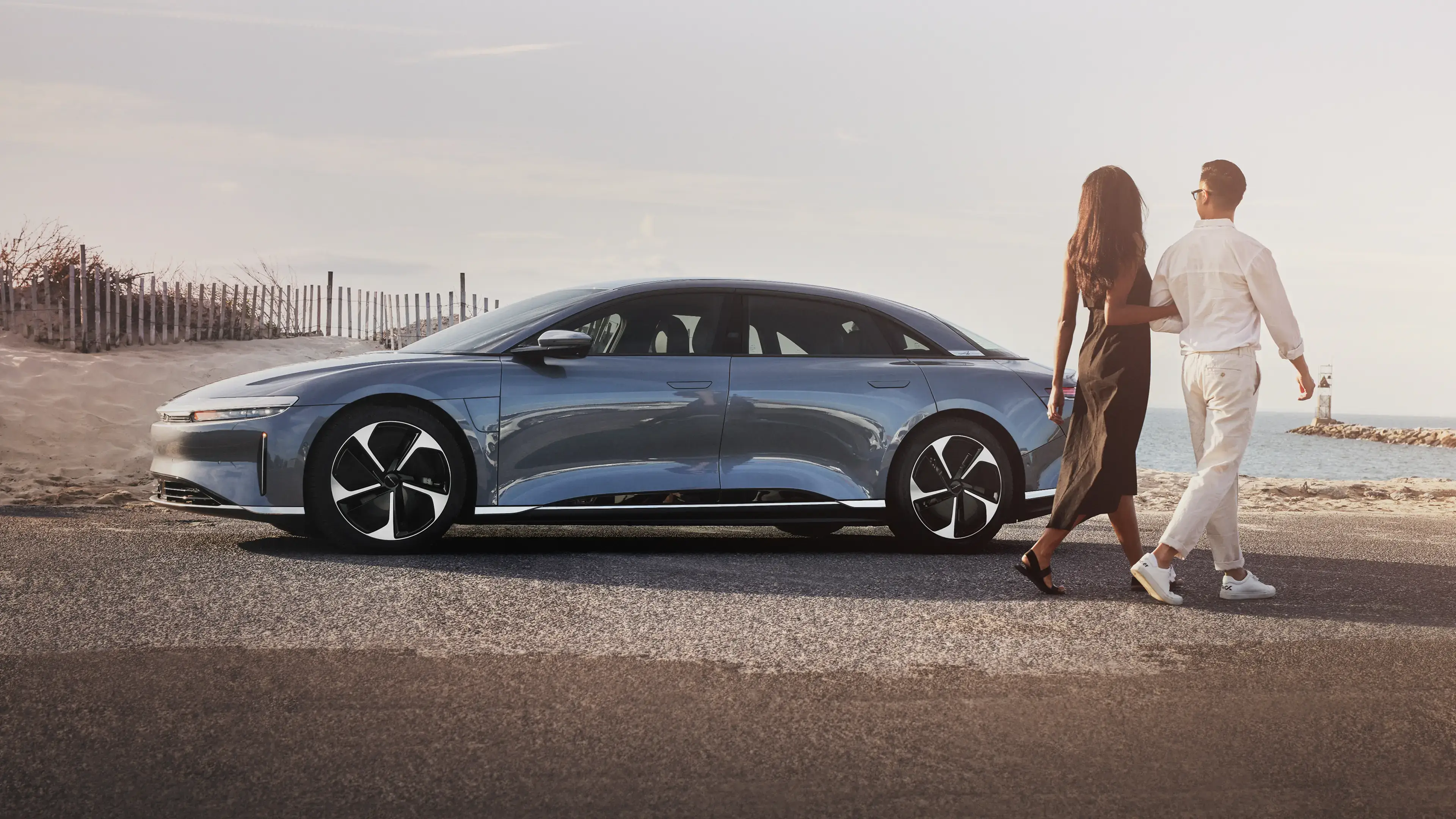 Couple walking past Lucid Air sedan parked on coastal road with ocean view at sunset