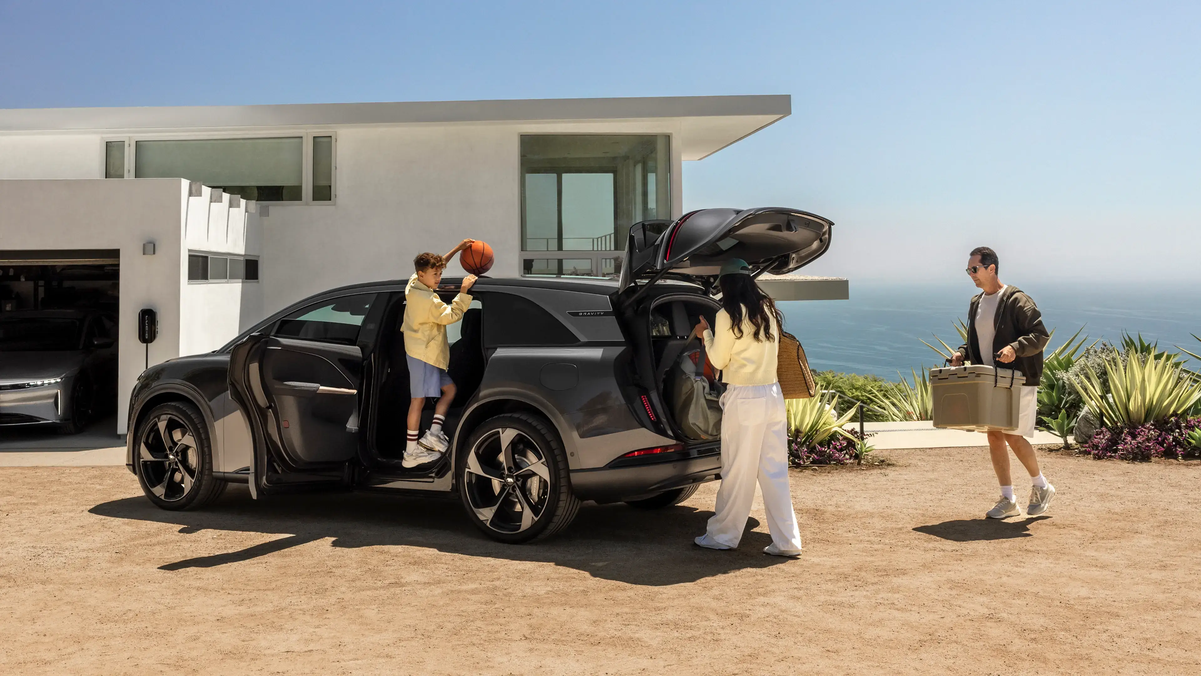 Family loading a dark gray SUV outside a modern coastal home; boy holds basketball, woman loads trunk, man carries a cooler.