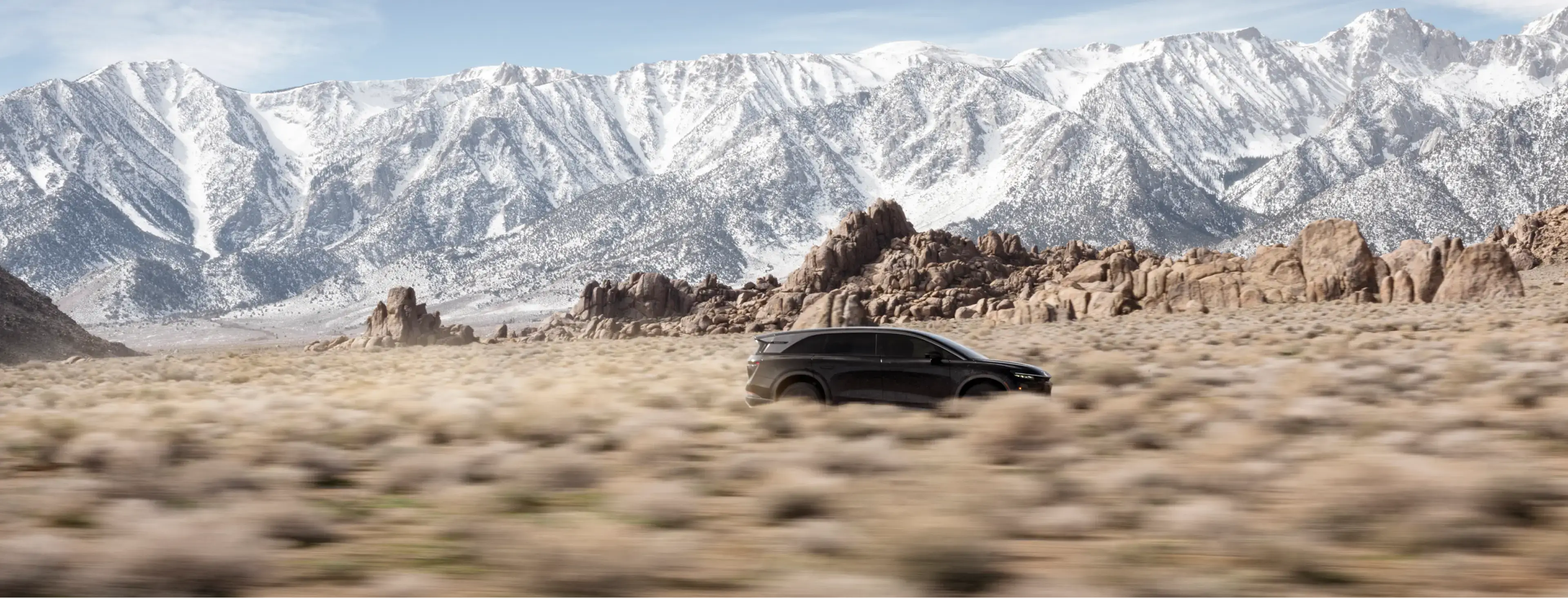 A black SUV drives through a desert landscape with rocky formations and snow-capped mountains in the background.