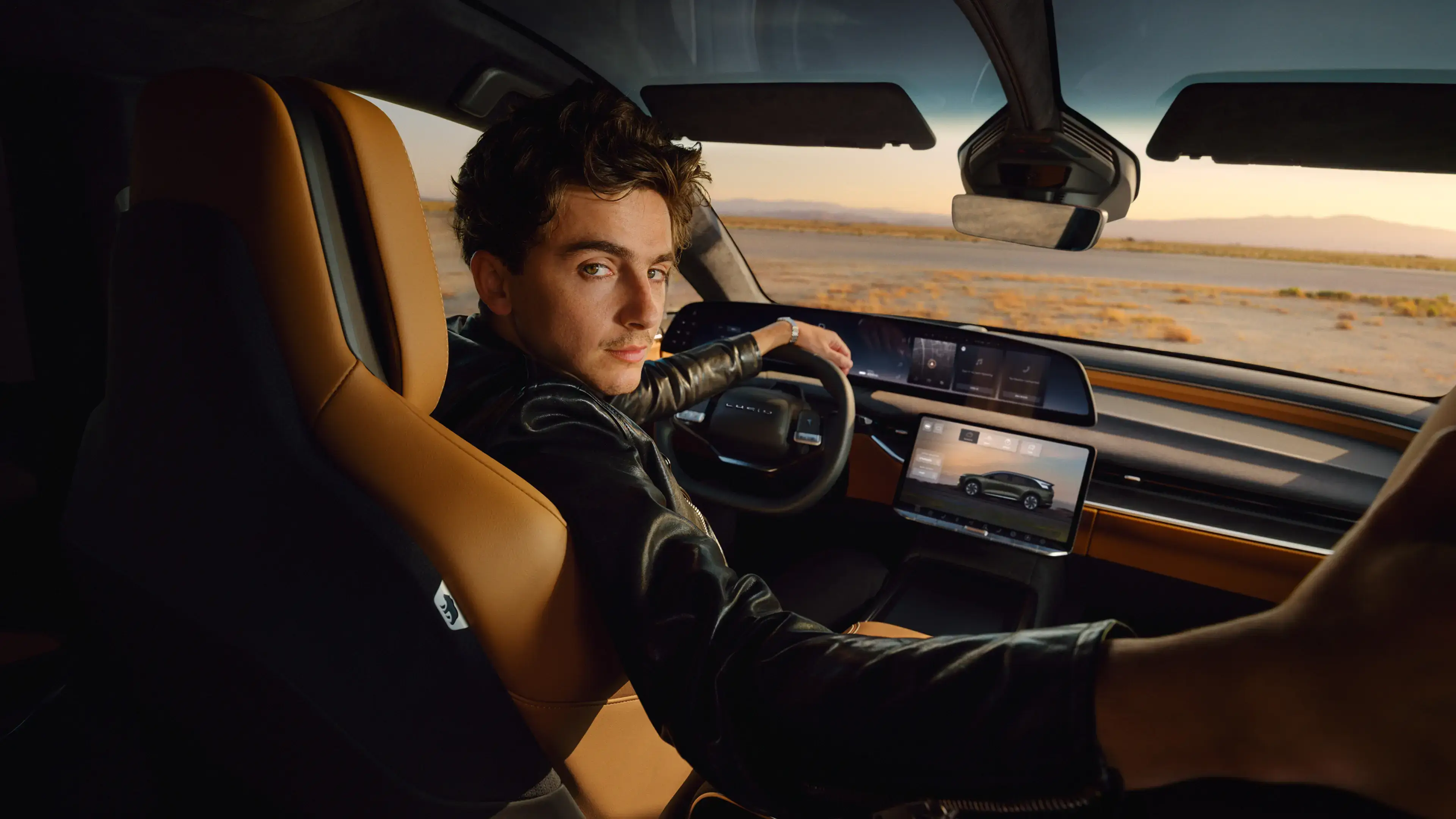 Timothèe Chalamet in leather jacket driving a Lucid Gravity with digital displays through a desert landscape at sunset