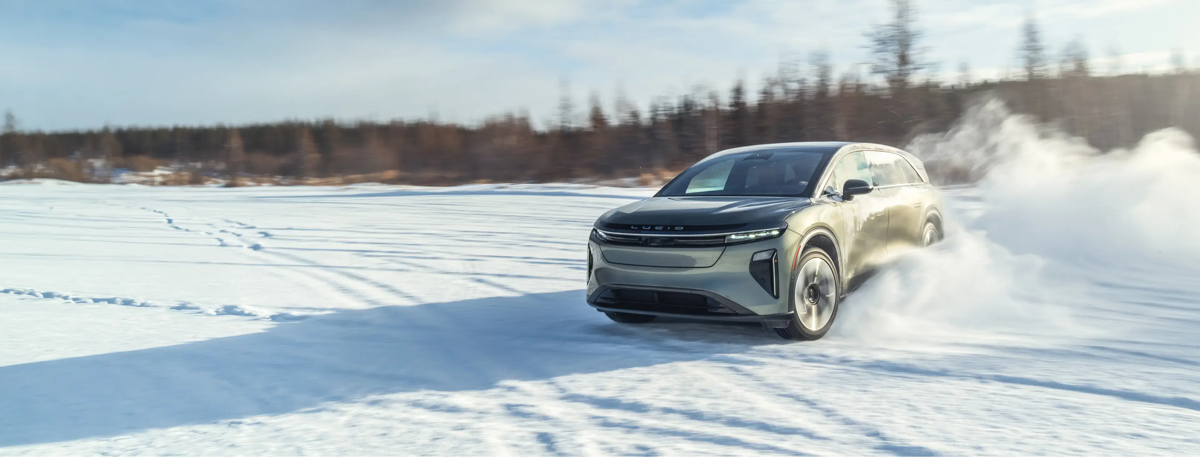 Lucid electric SUV driving across snowy terrain, kicking up powder with forest backdrop under blue sky.