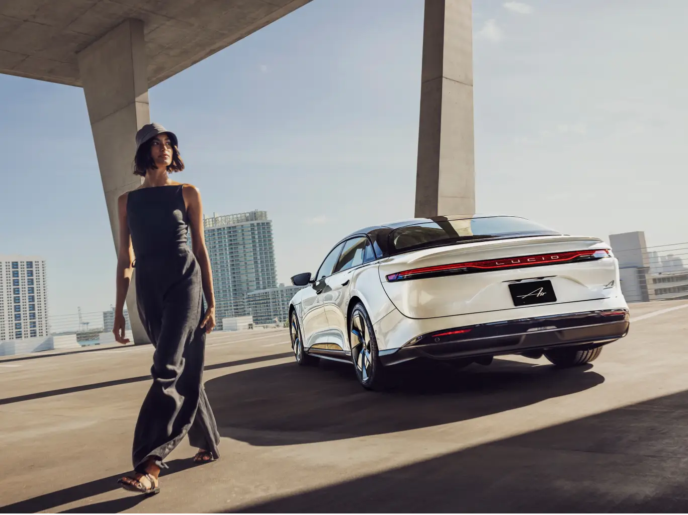 Person in black outfit walking beside a Lucid Air under modern concrete architecture with city skyline.