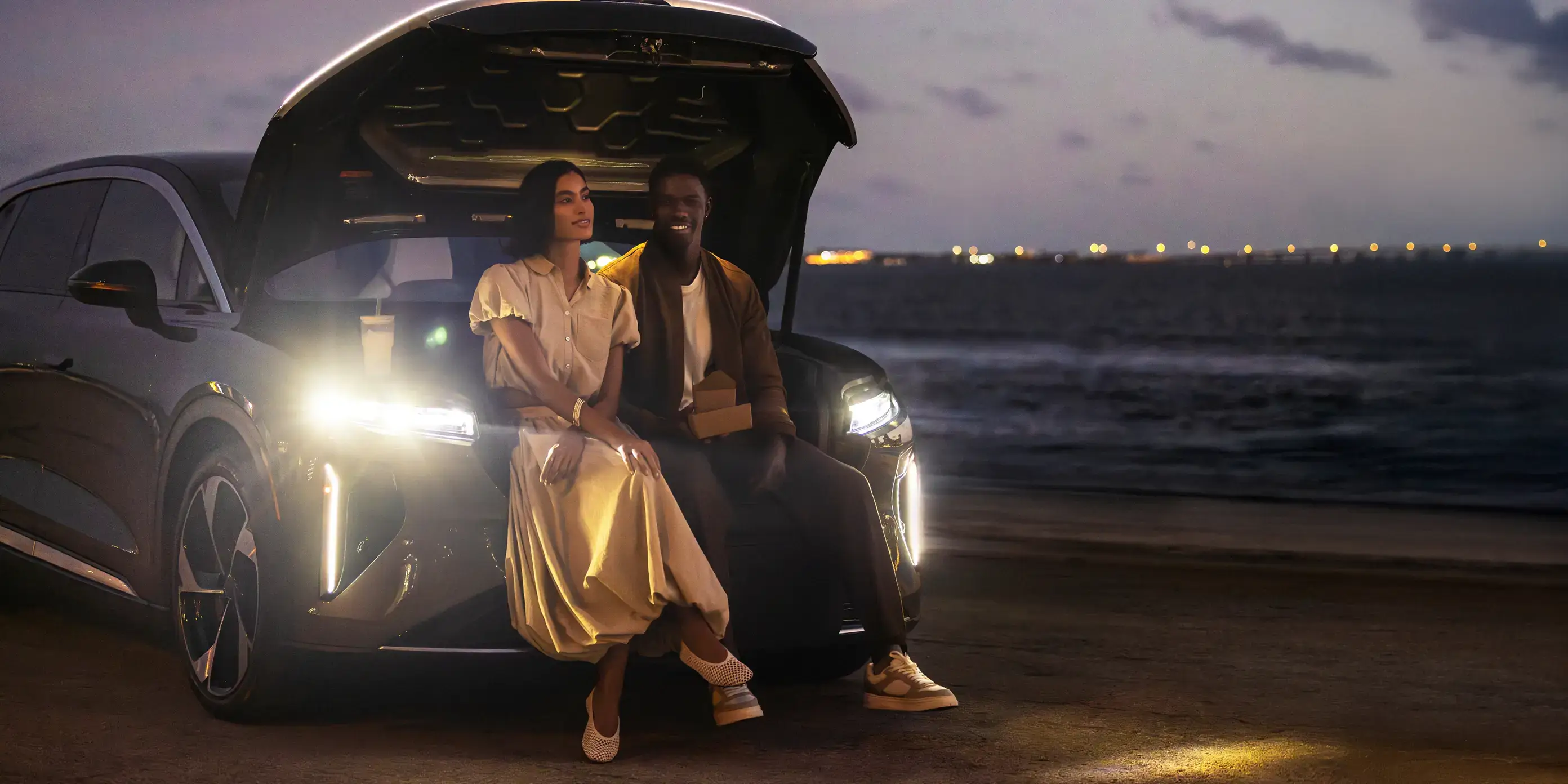 Two people sitting on the trunk of a Lucid Gravity at twilight by the ocean, city lights glowing in the distance.