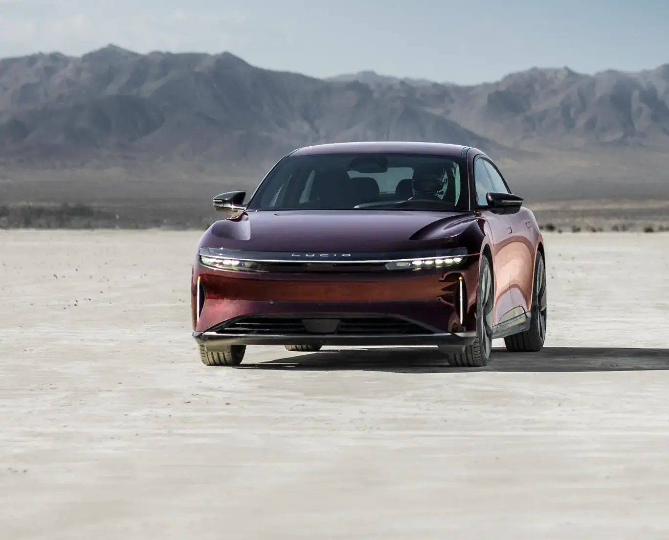 A deep red Lucid Air electric sedan on a desert salt flat, with mountains and a hazy sky in the background.