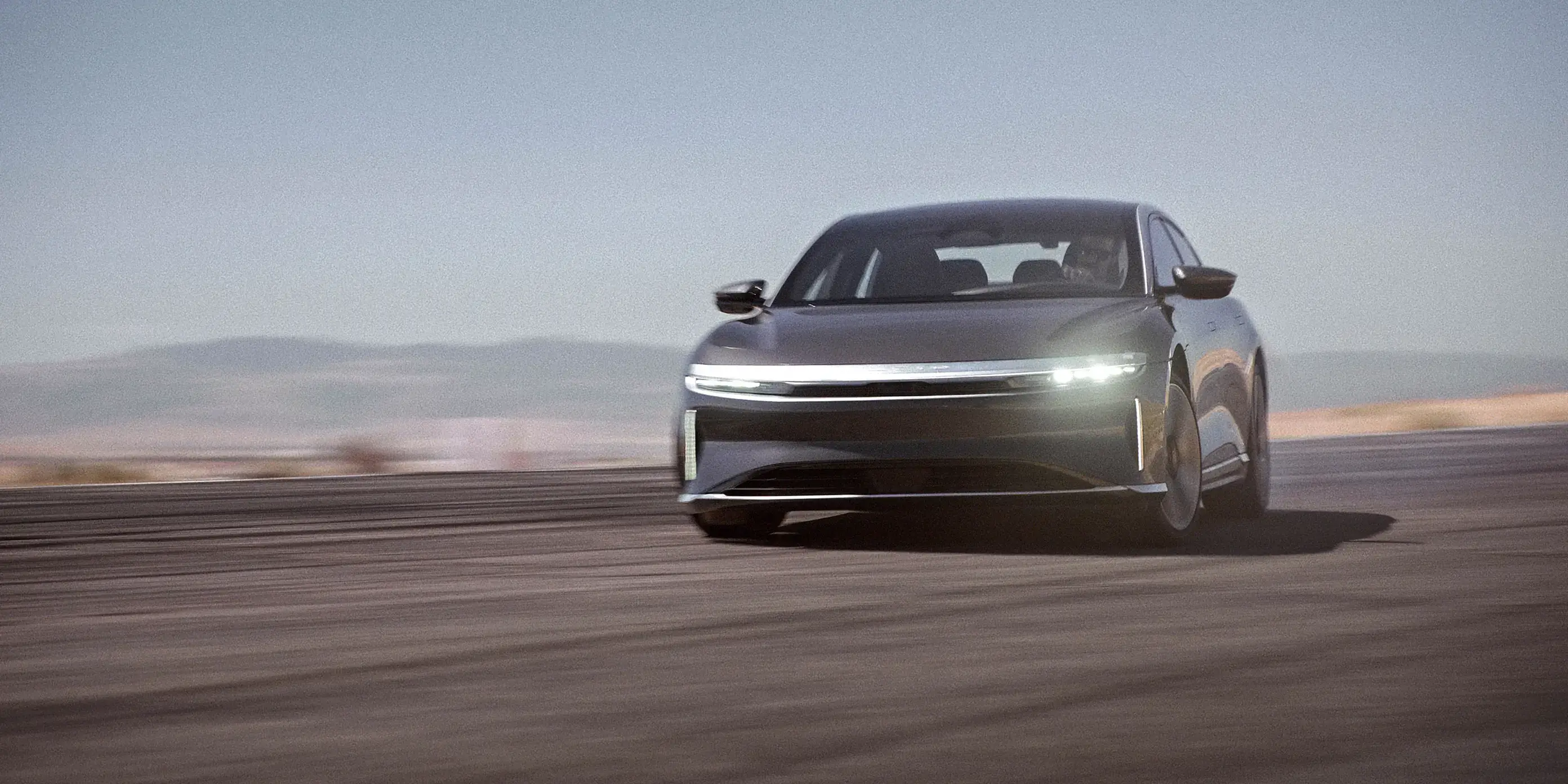 Berline Lucid Air roulant sur une route désertique avec les phares allumés, montagnes visibles en arrière-plan sous un ciel gris.