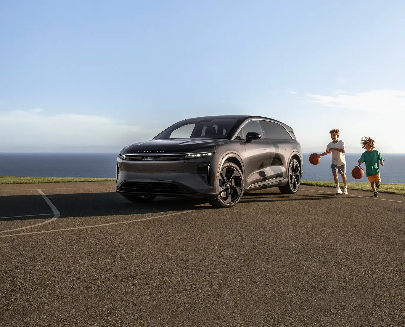 A gray Lucid SUV parked on an outdoor basketball court near the ocean, with two children dribbling basketballs nearby.