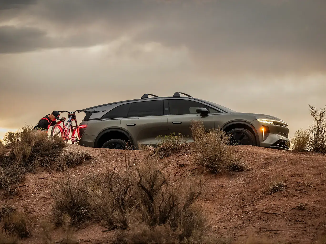 Lucid Gravity SUV parked on a red dirt desert hill with a person unloading a red bicycle from the rear hitch rack.