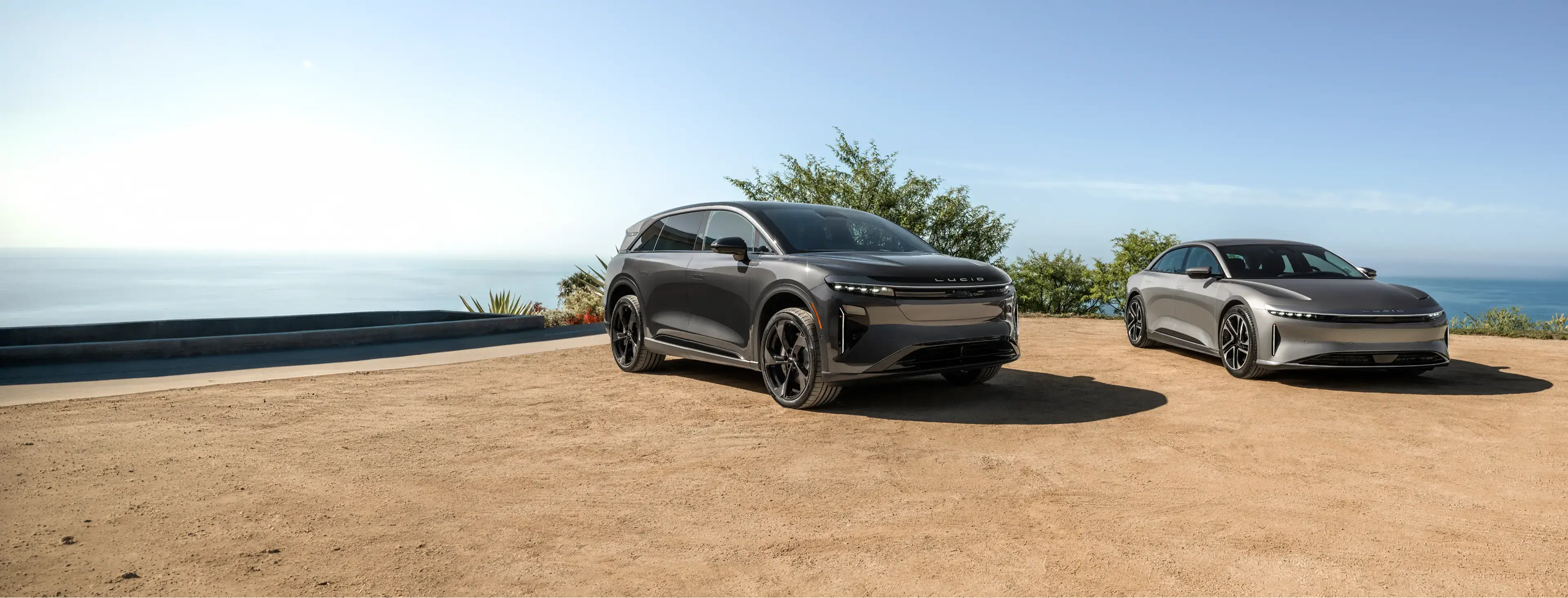 Two Lucid electric vehicles, a dark gray SUV and a silver sedan, parked on a sandy overlook with ocean and blue sky.