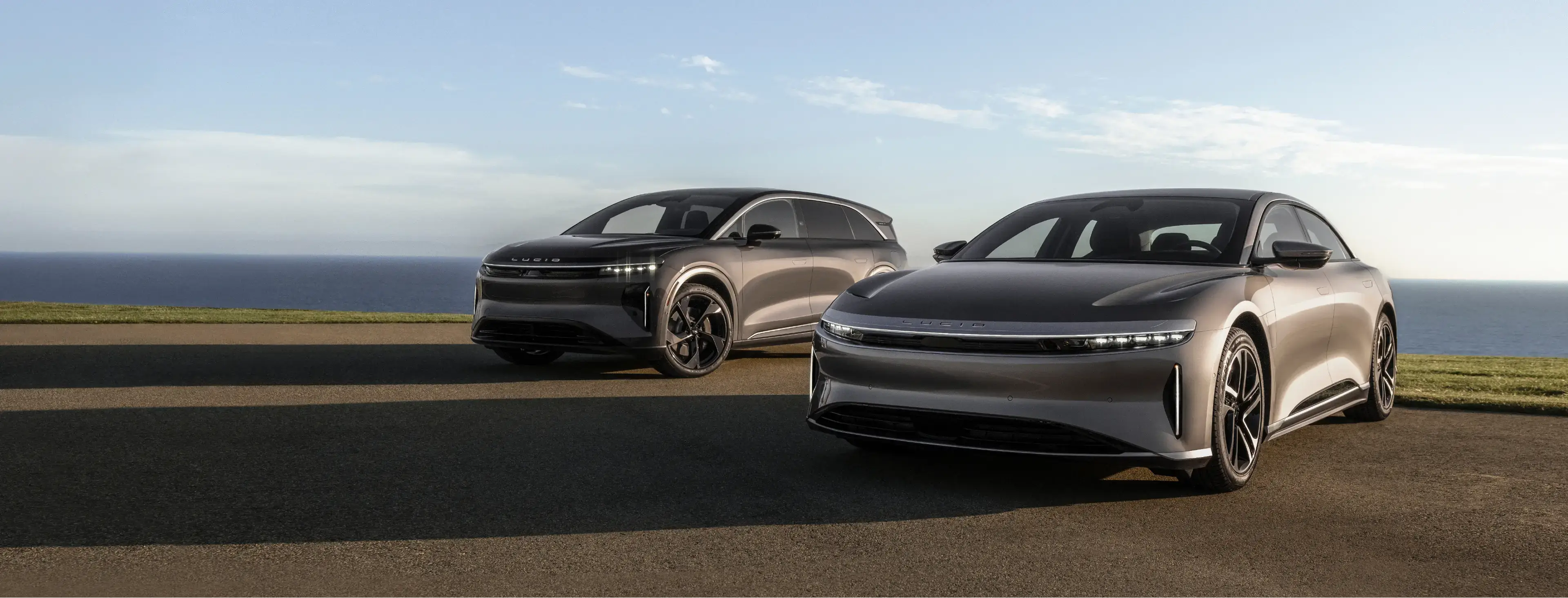 Two silver Lucid electric vehicles parked near a coastal overlook with green grass, ocean, and blue sky in the background.