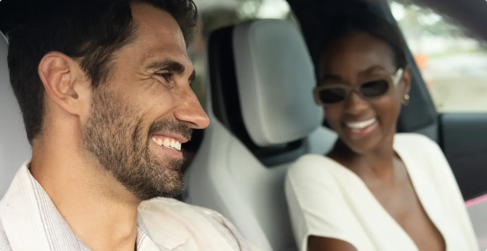 Deux personnes souriantes à l’intérieur d’une voiture : un homme à l’avant et une femme portant des lunettes de soleil sur la banquette arrière.