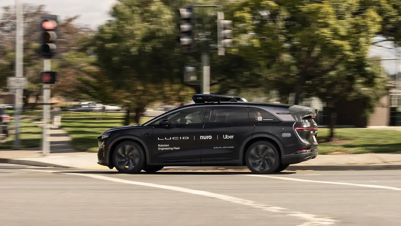Schwarzes autonomes Fahrzeug von Lucid mit Uber- und Nuro-Branding fährt über eine Kreuzung mit Ampeln und Bäumen.