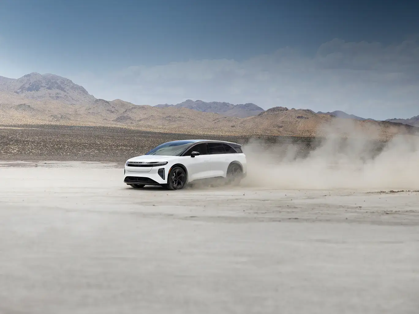White electric SUV driving across a dusty desert salt flat, kicking up dust, with mountains in the background.