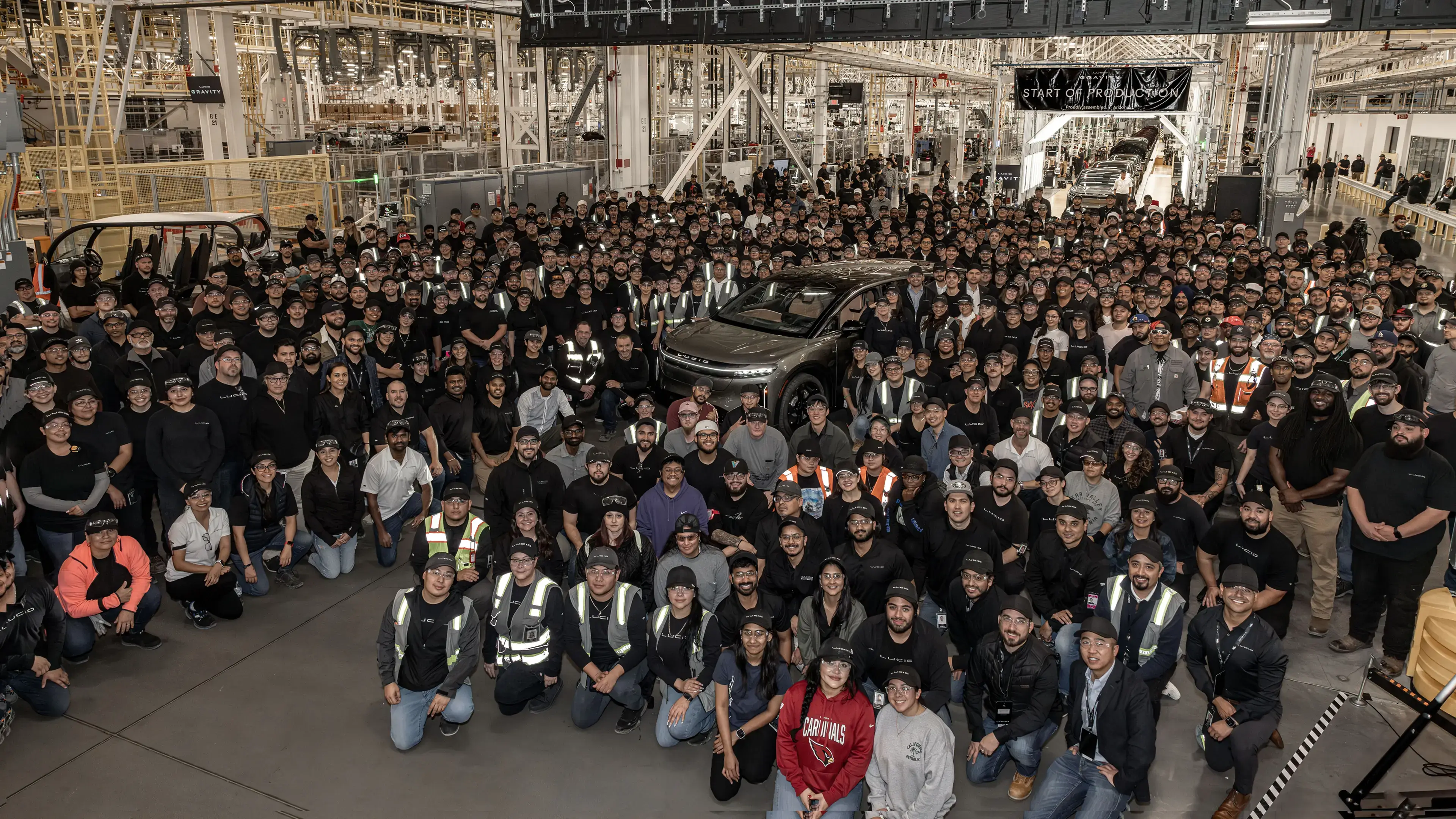 Large group of automotive factory workers gathered around a new electric vehicle at a production facility launch event.