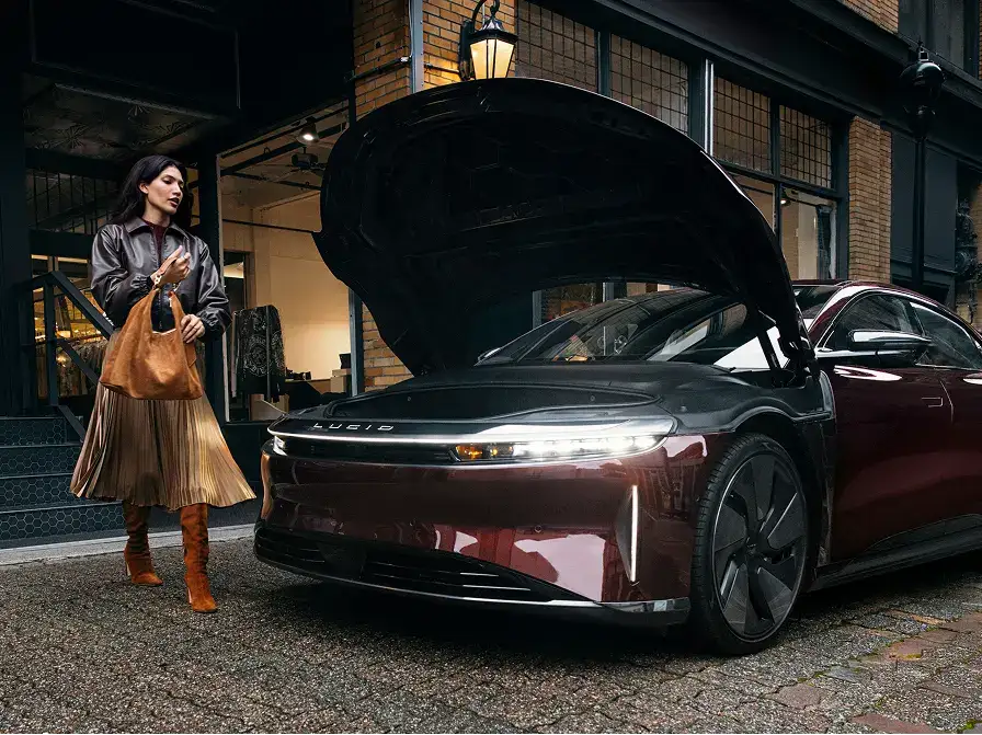 Woman in leather jacket and pleated skirt approaching a burgundy Lucid electric car with open hood outside a brick building.