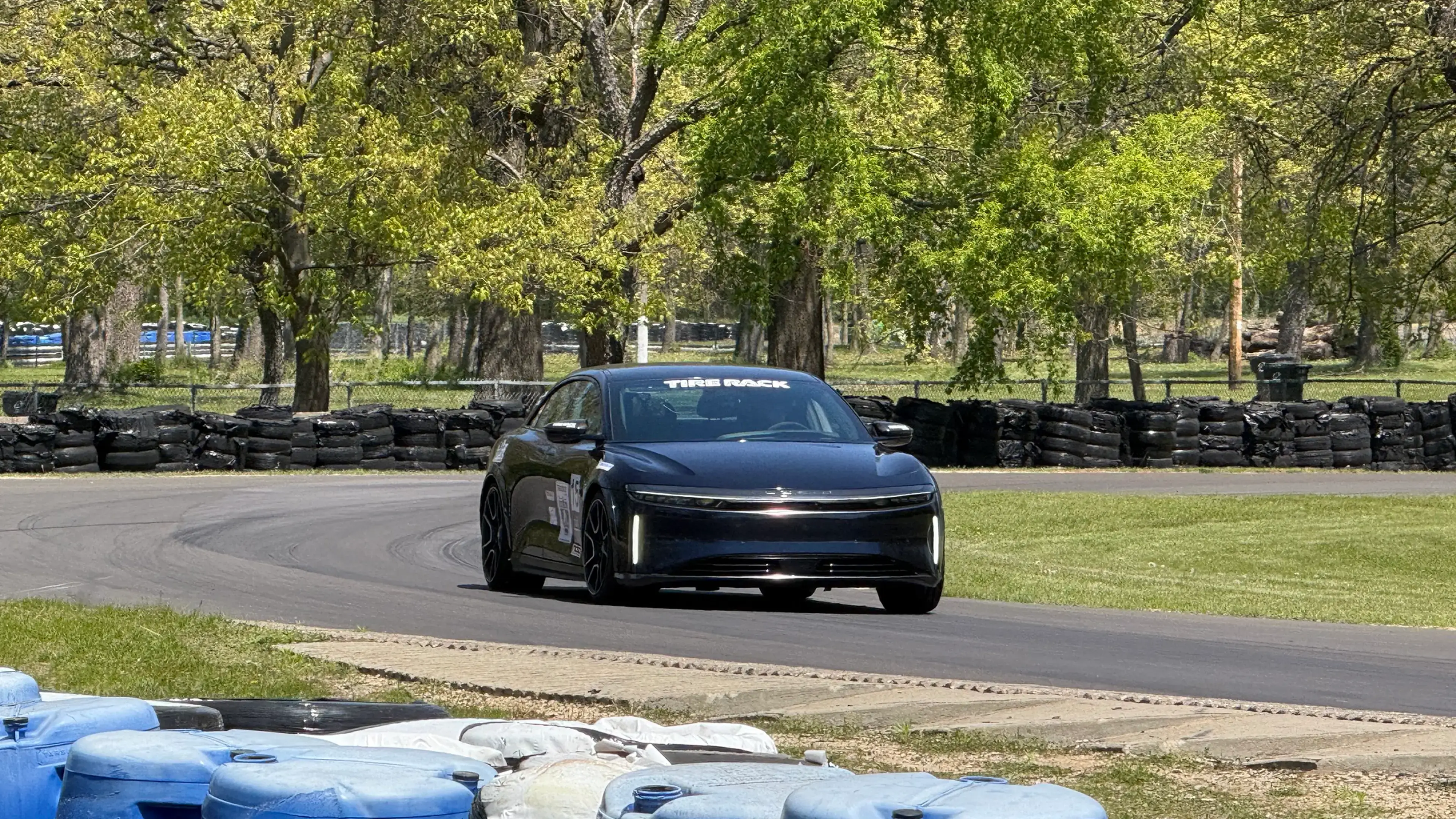 A Lucid Air Sapphire sedan navigates a curve on a race track, bordered by tire walls and blue barrels, with green trees behind.