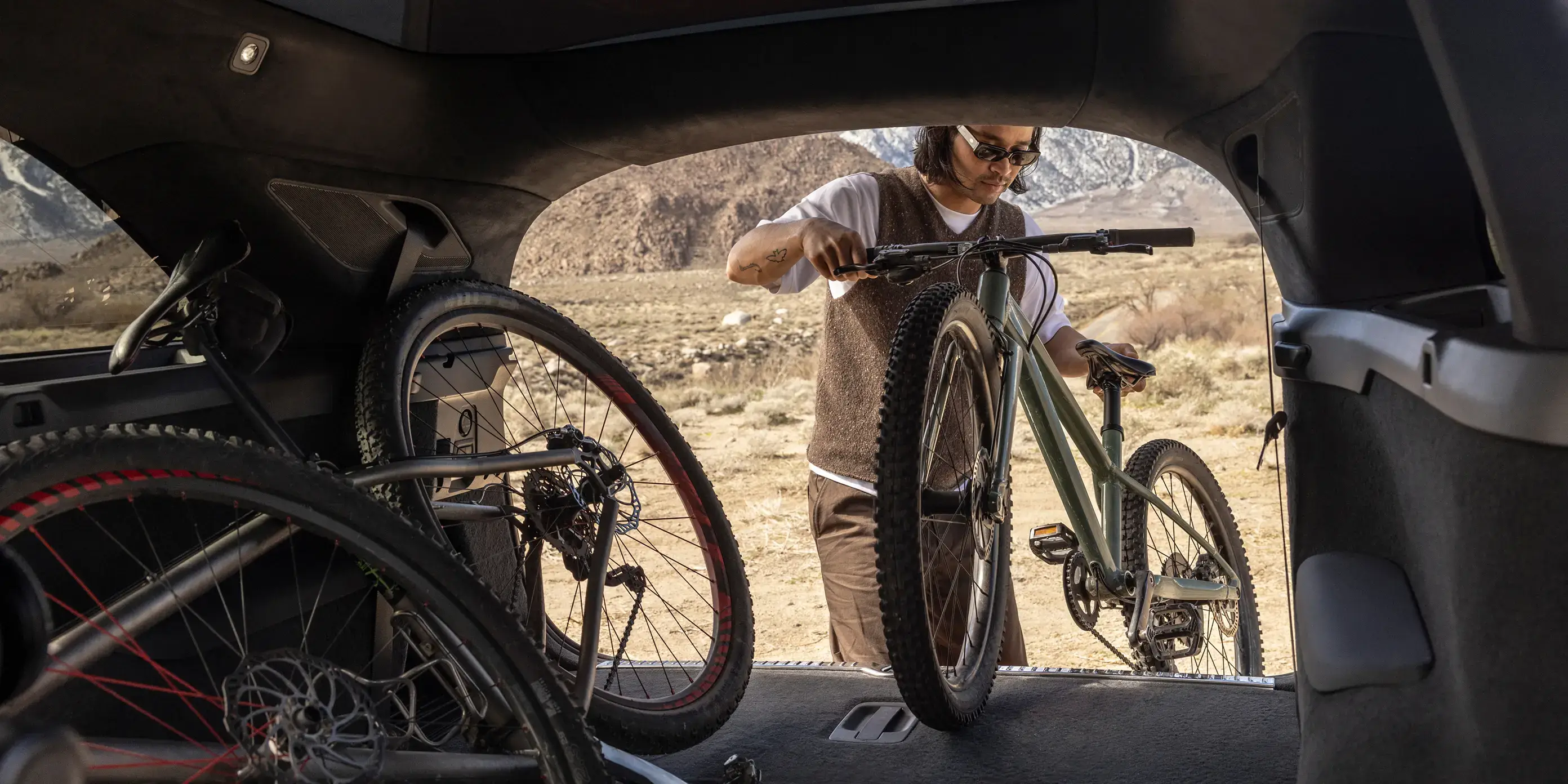 View from inside a vehicle as a person loads a mountain bike into the trunk, with desert landscape in background.
