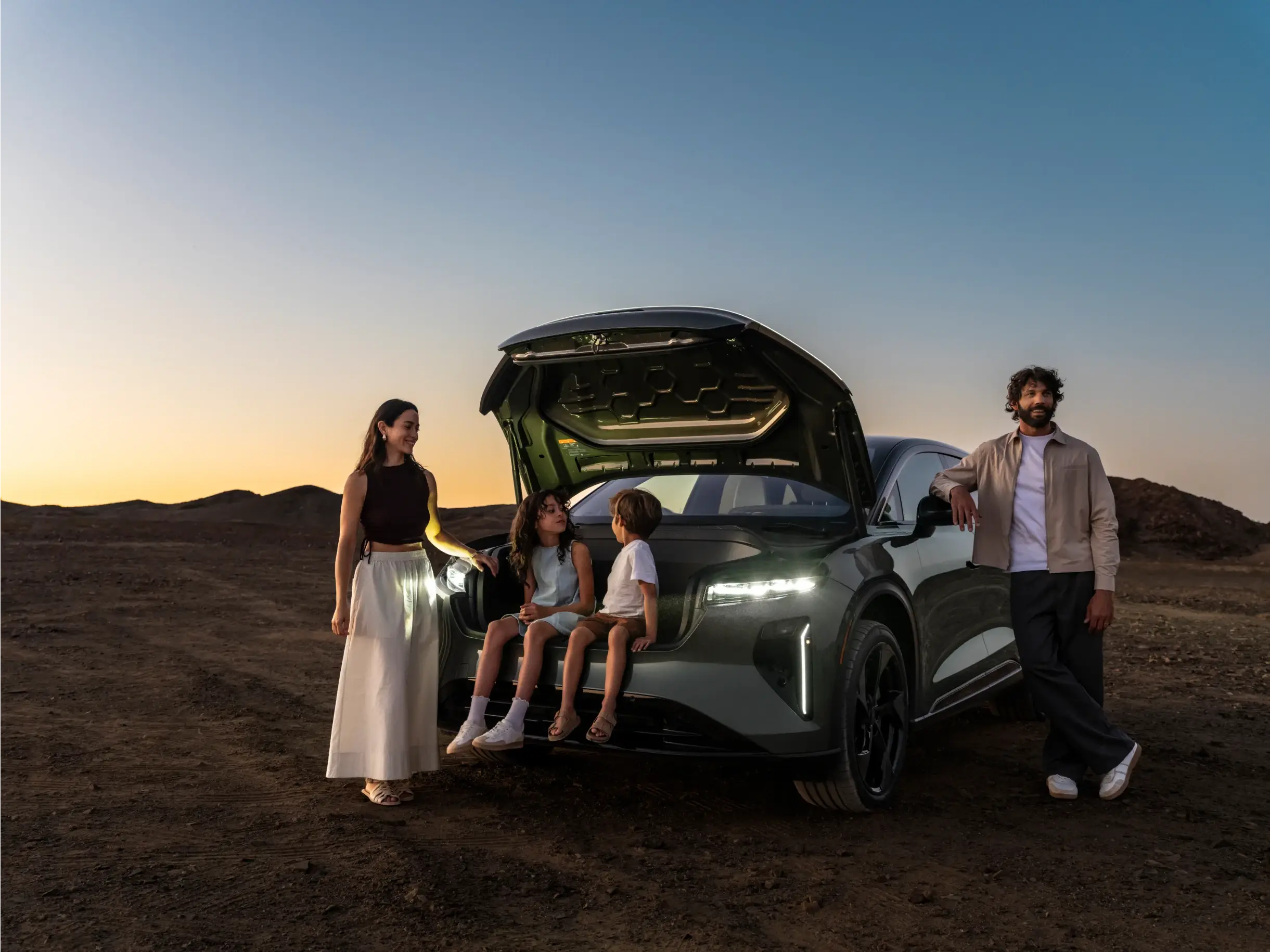 Eine Familie versammelt sich bei Sonnenuntergang um eine Lucid Gravity, wobei zwei Kinder im geöffneten Kofferraum sitzen und die Erwachsenen daneben in der Wüstenlandschaft stehen.