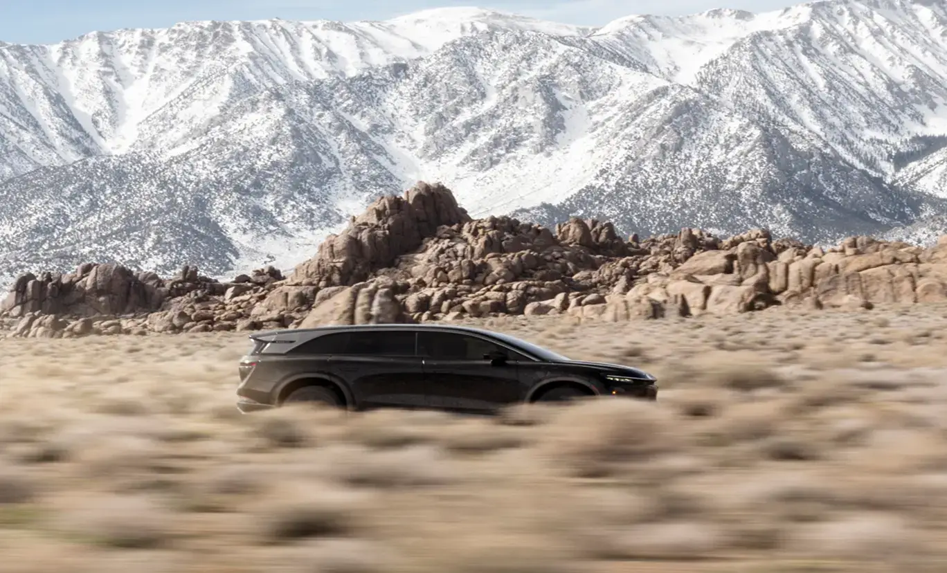 A Lucid Gravity SUV speeds across a desert landscape with rugged rock formations and snow-capped mountains in the background.