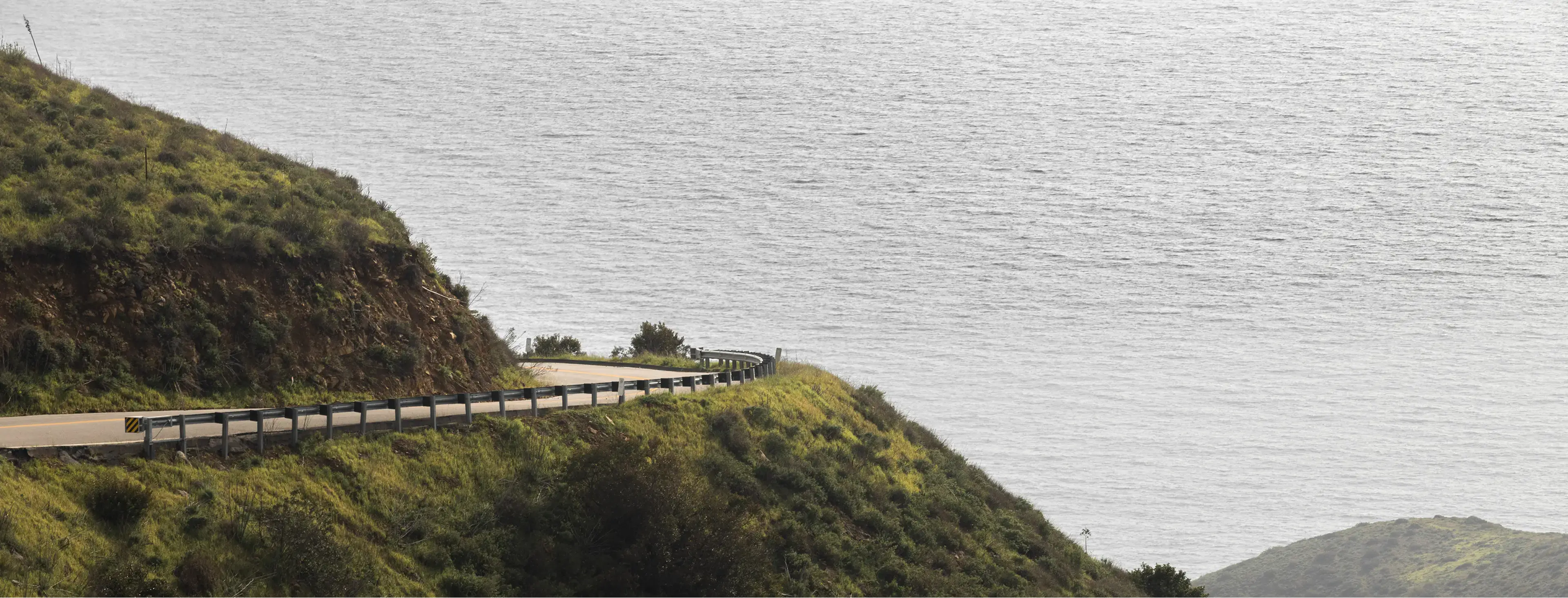Winding coastal road curves along green hillside cliffs overlooking calm gray ocean waters.