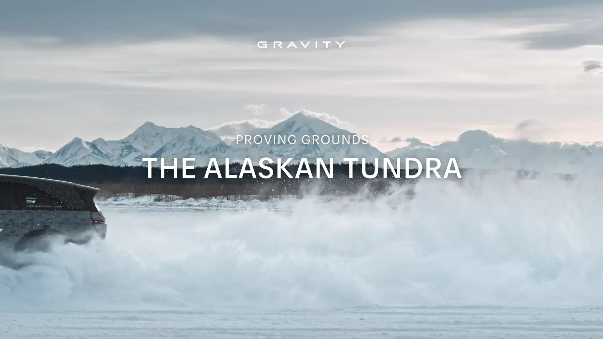 Vehicle driving through snowy Alaskan tundra with snow-capped mountains in background and "GRAVITY" branding visible.