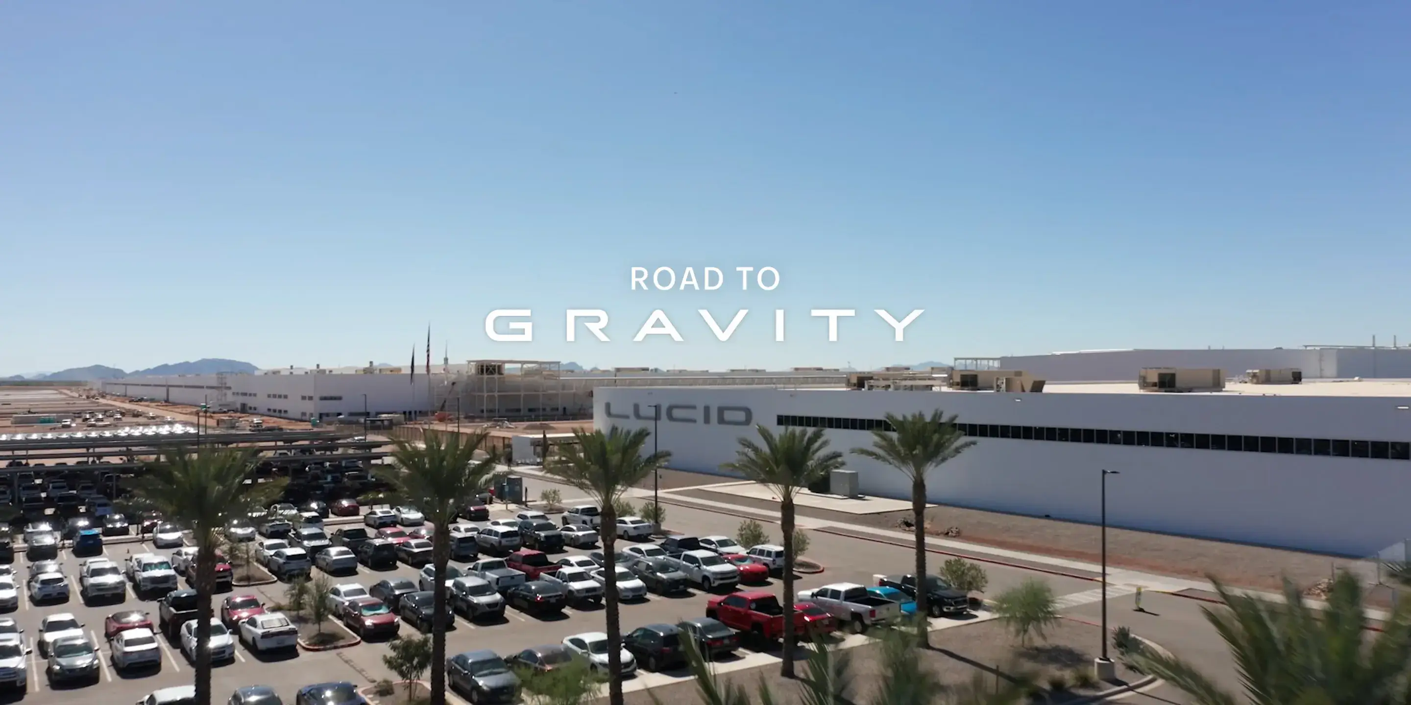 Lucid Motors facility with "ROAD TO GRAVITY" text overlay, showing white buildings and a full parking lot under blue sky.