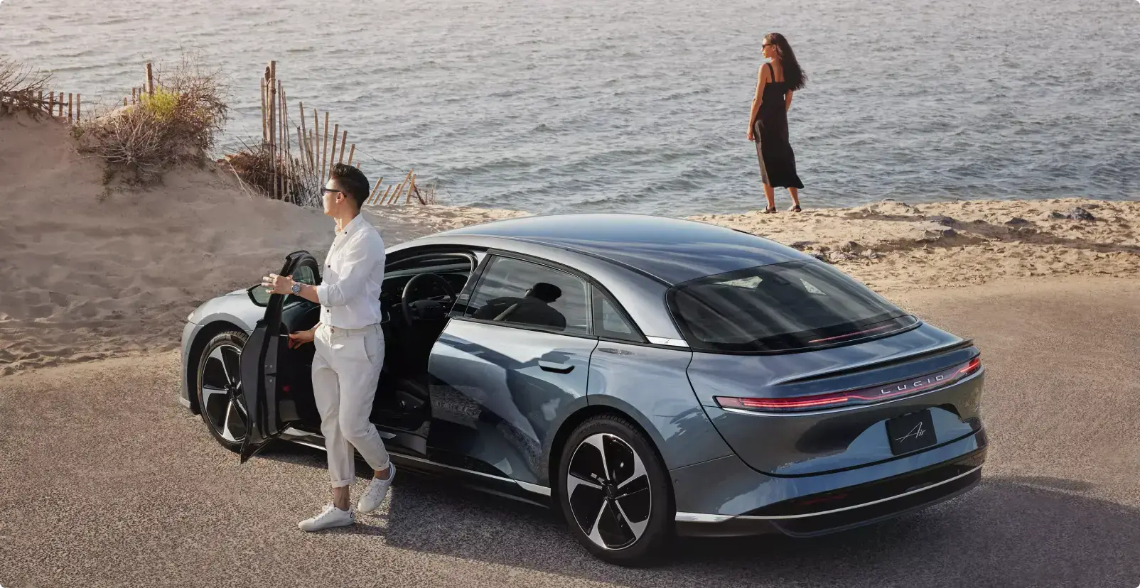 A man in white opens a blue Lucid Air sedan near a beach while a woman in a black dress stands by the ocean.