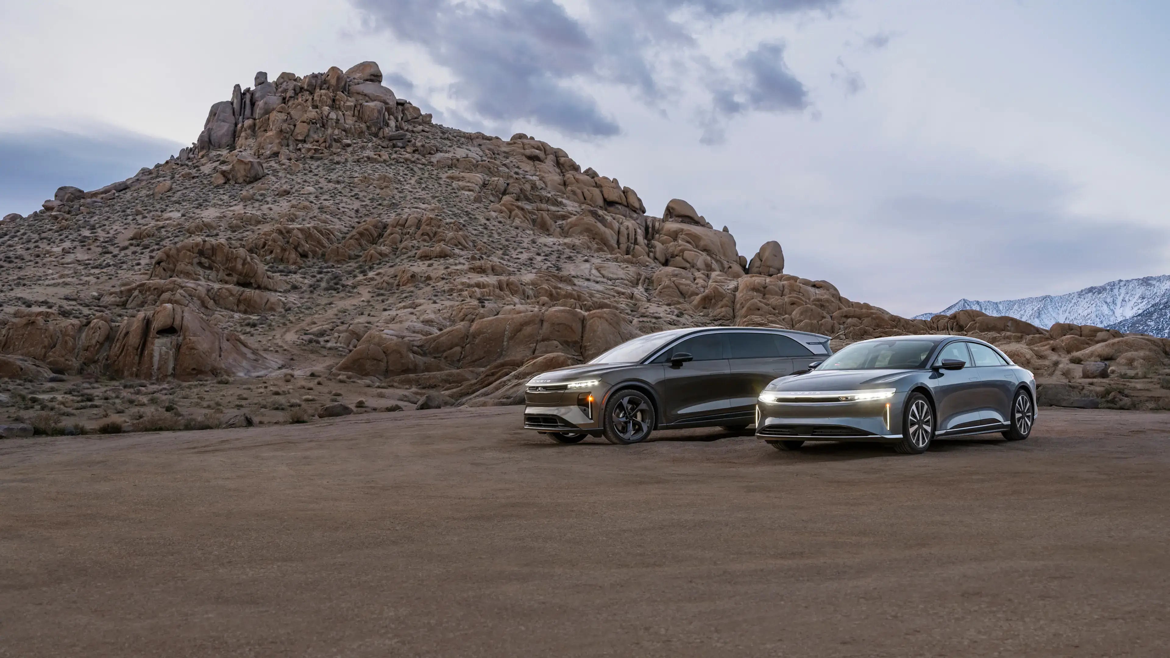 Two Lucid electric vehicles parked in a rocky desert landscape with snow-capped mountains and a cloudy sky at dusk.