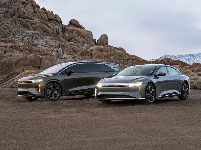 Two sleek Lucid electric vehicles parked on rocky desert terrain, headlights on, with rugged boulders and mountains behind them.