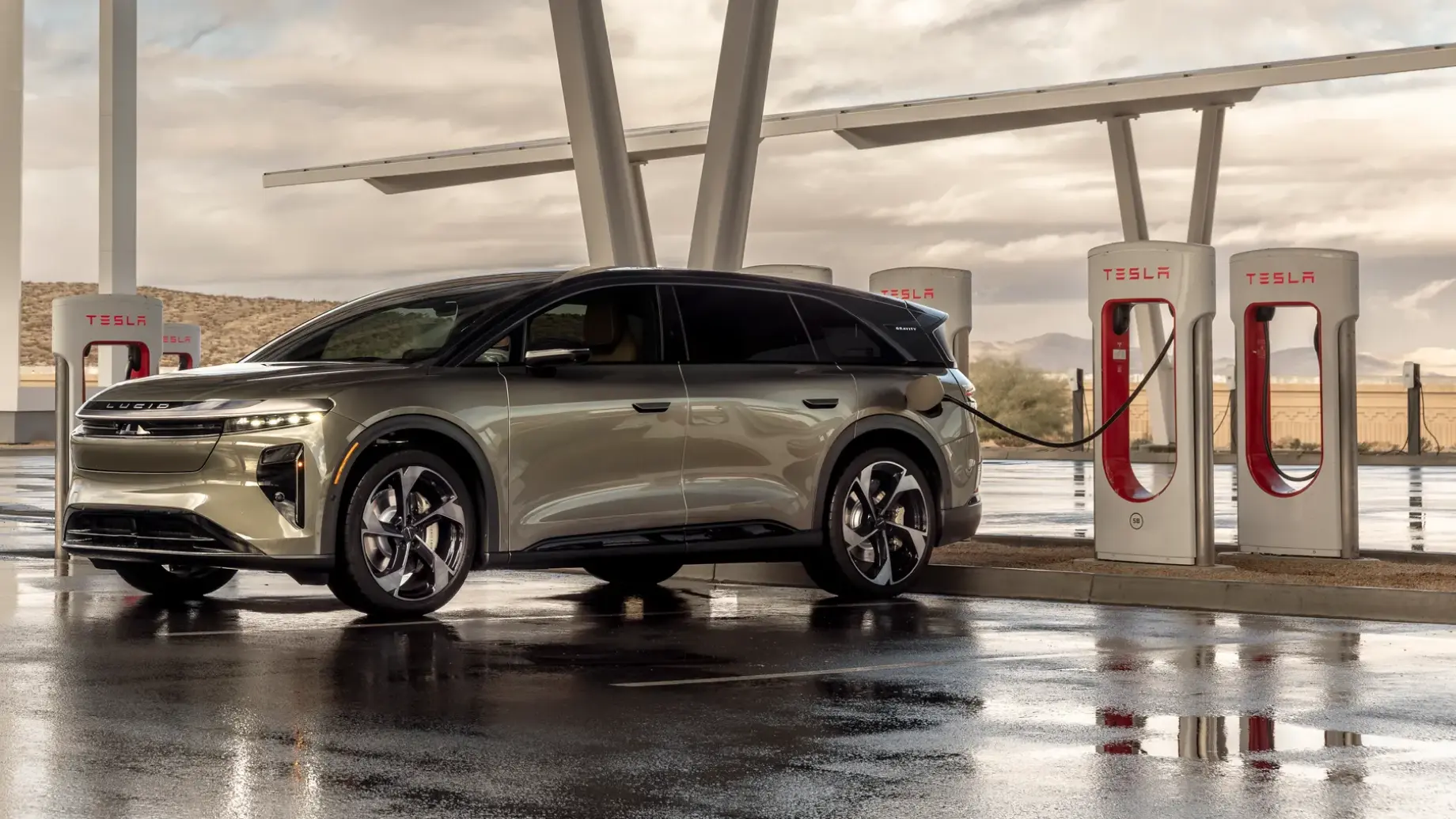 Lucid Gravity charging at a Tesla Supercharger station under a covered canopy on a cloudy day.