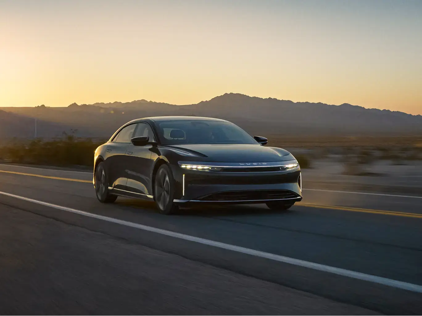 A dark gray Lucid Air electric sedan driving on a desert highway at sunset with mountains in the background.