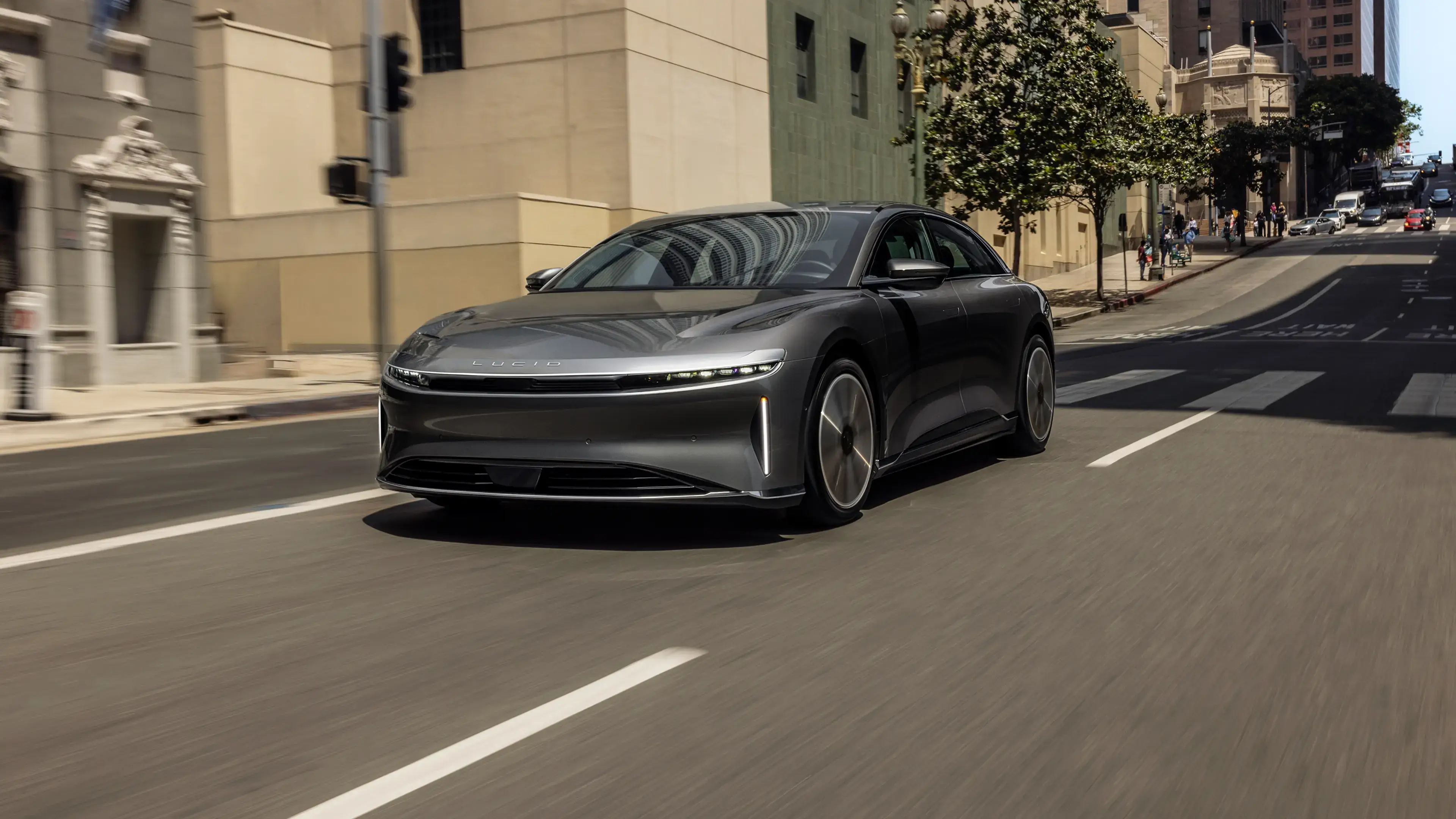 A sleek gray Lucid Air electric sedan driving on a city street, with modern buildings in the background.