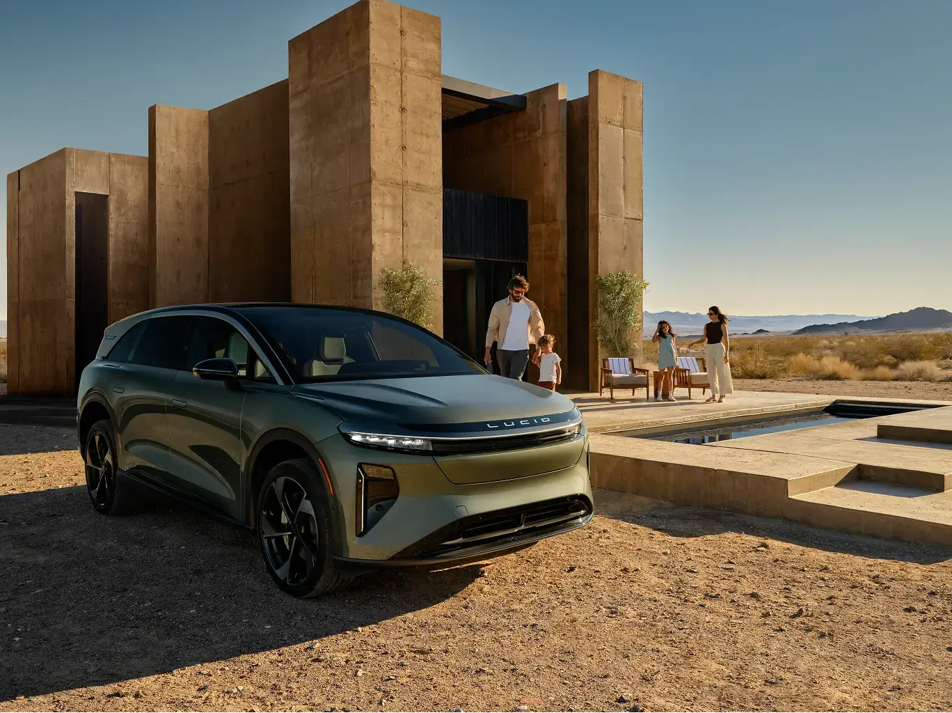Lucid electric SUV parked outside modern desert home with family gathering on patio, mountains in background at sunset.