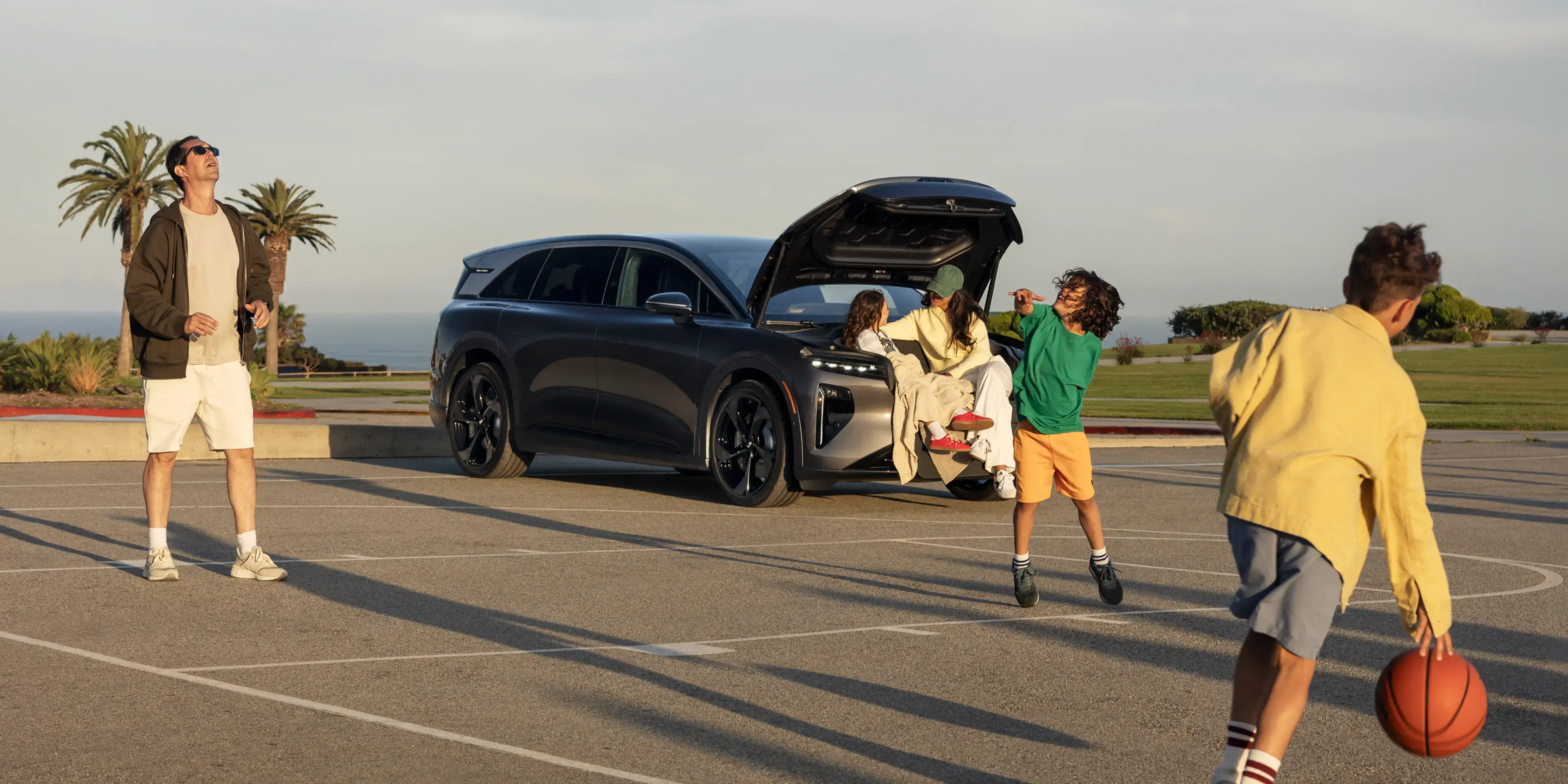 Family playing basketball in a parking lot near a dark gray SUV with open trunk, palm trees and ocean in background.