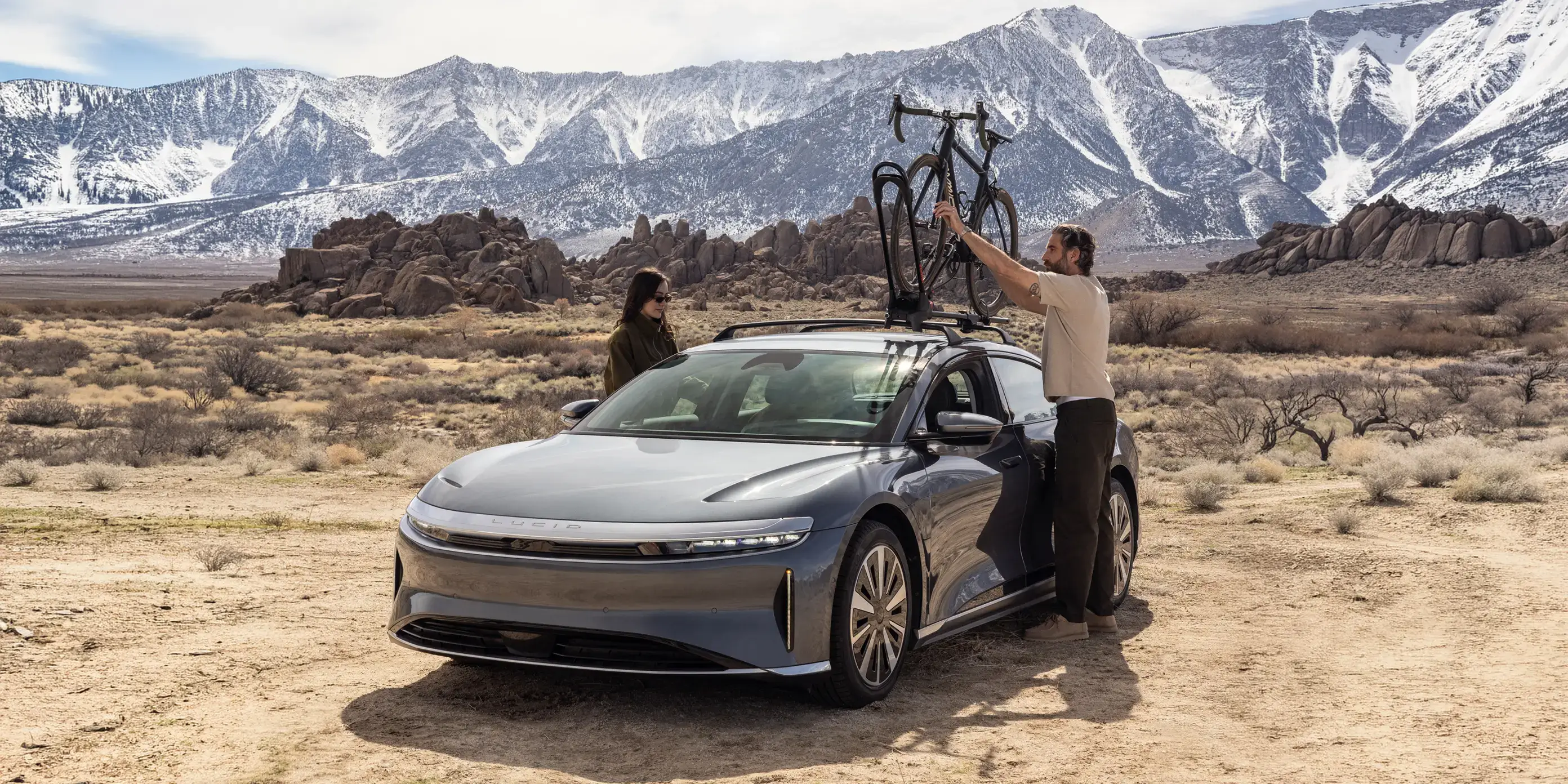 Person removing bicycle from car roof rack in desert landscape with snow-capped mountains in background.