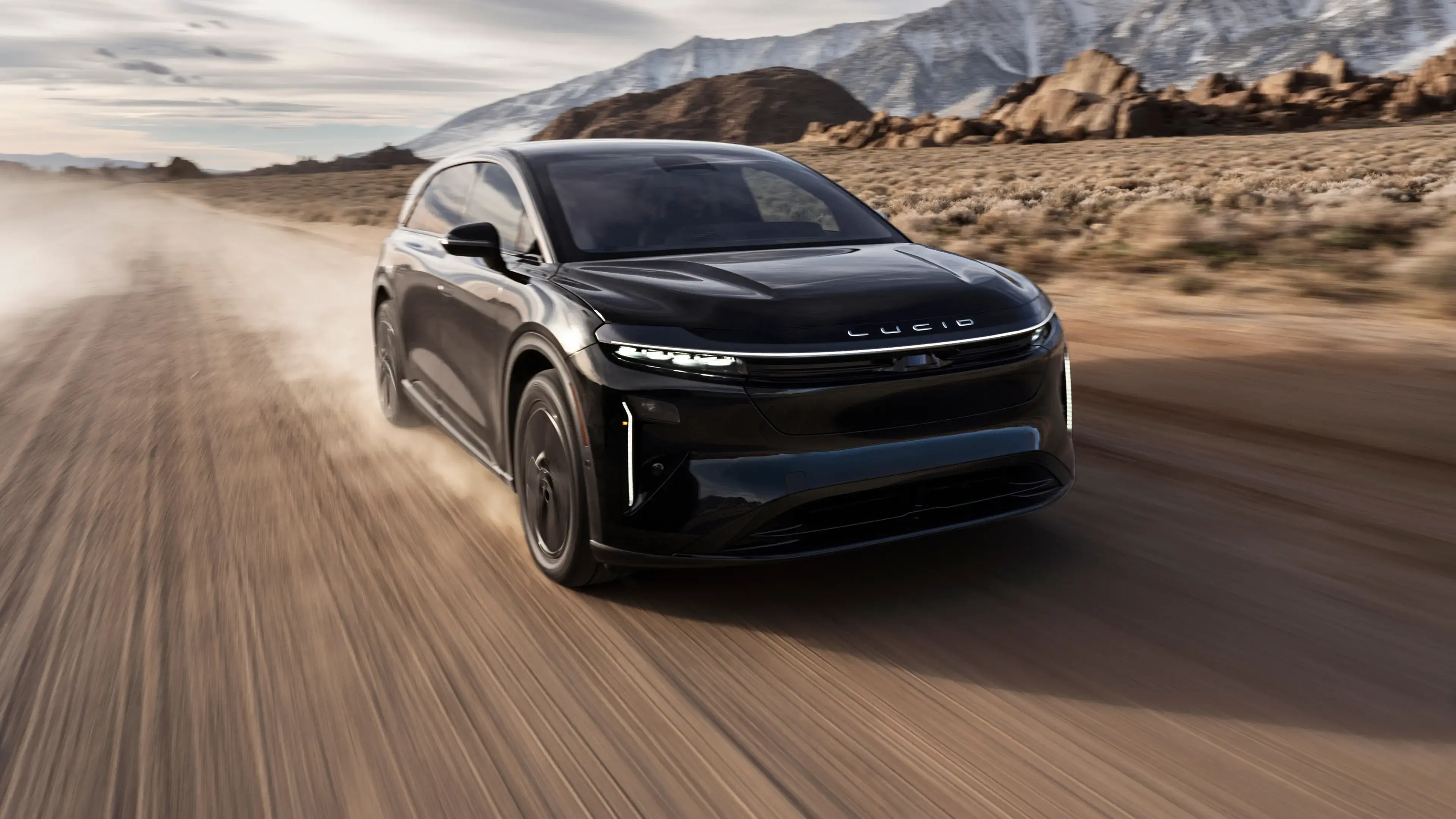 A black Lucid electric SUV speeding along a desert road with rocky, snow-capped mountains in the background.