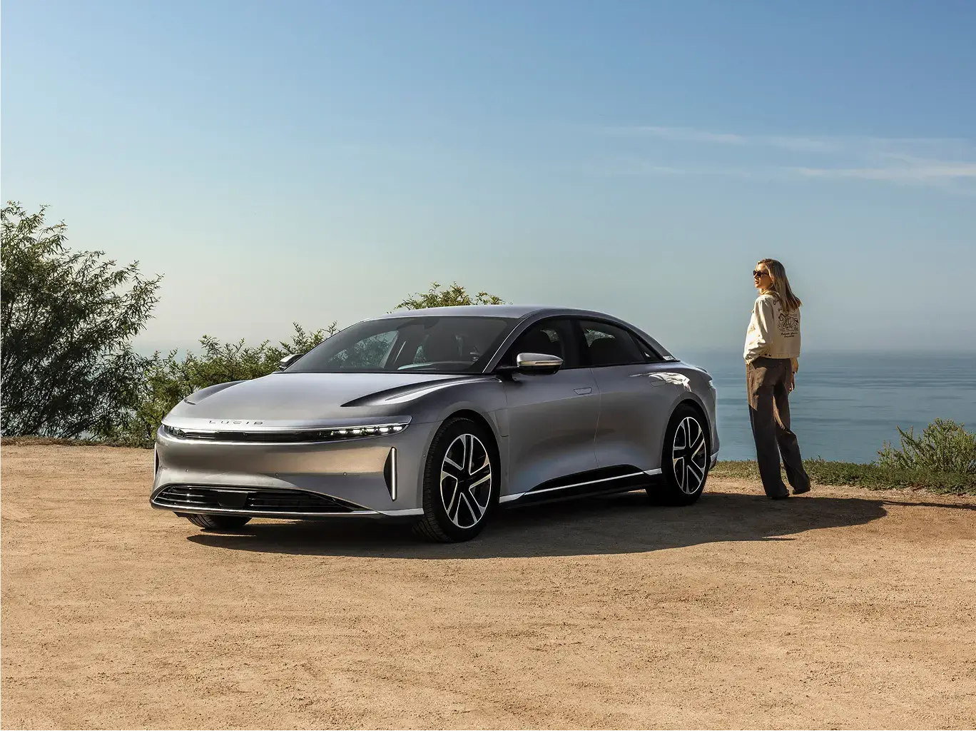 A Lucid Air electric sedan parked on a coastal overlook, with a woman standing beside it under a clear blue sky.