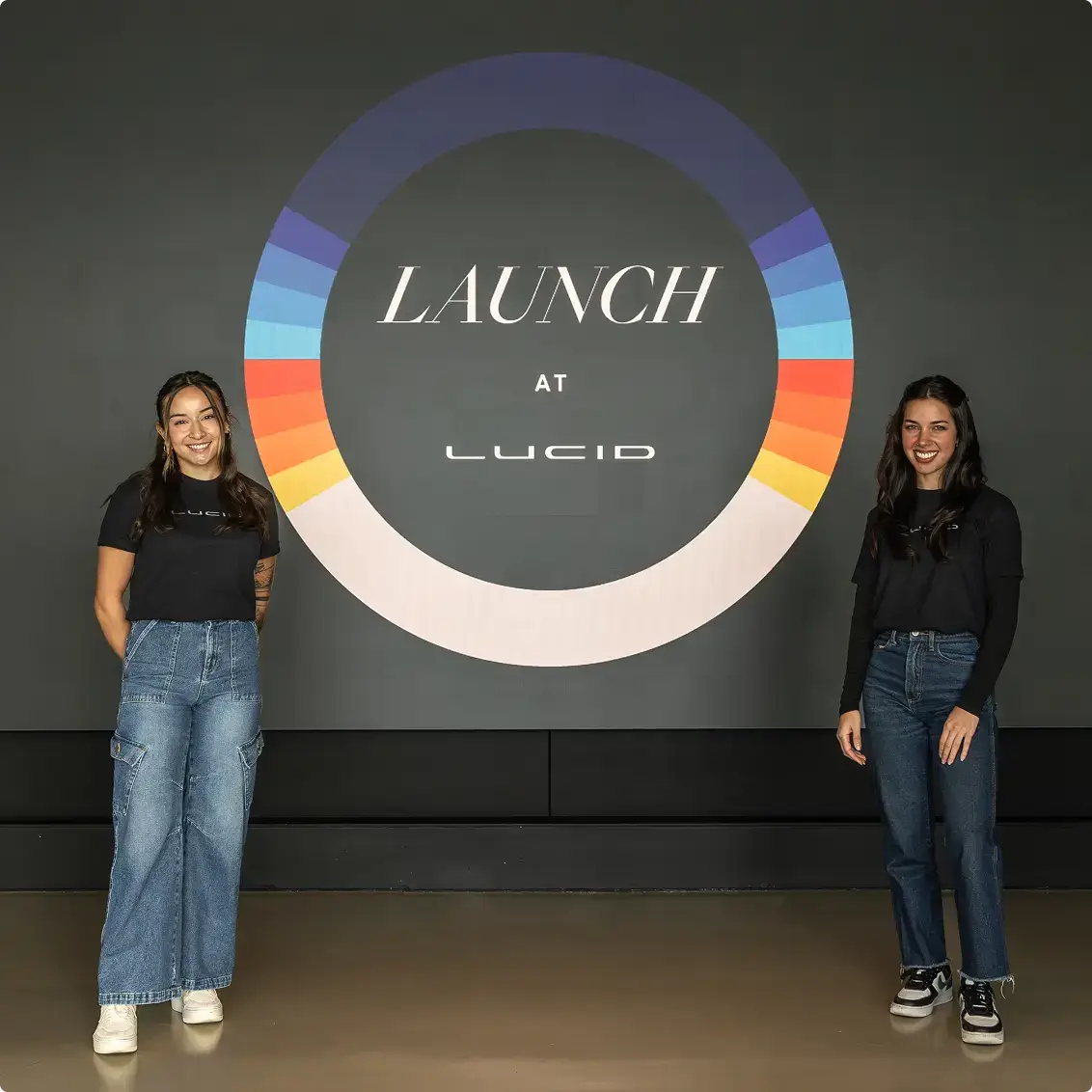 Two people in Lucid branded shirts standing beside a colorful circular "LAUNCH at LUCID" logo display on a dark wall.