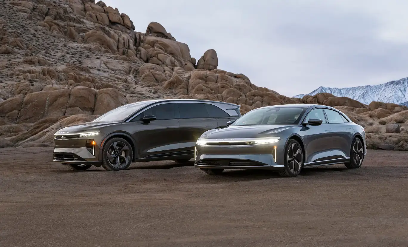 Two sleek Lucid electric vehicles parked in a desert landscape with rocky formations and snow-capped mountains in the background.