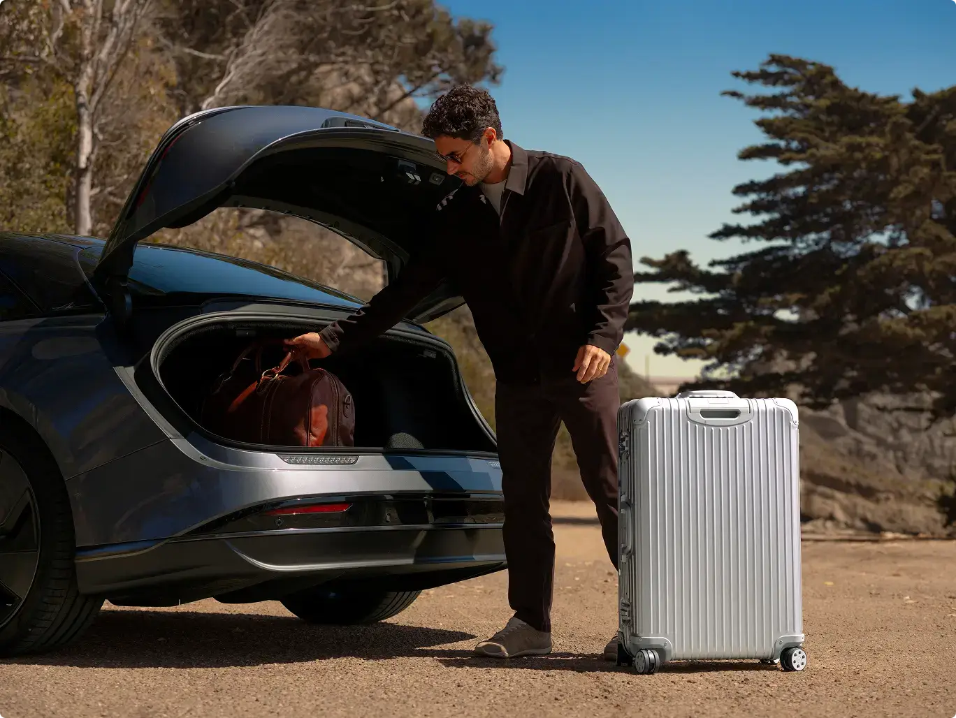 A man in dark clothing loads luggage into a Lucid Air trunk, with a silver hard-shell suitcase standing nearby on a sunny day.